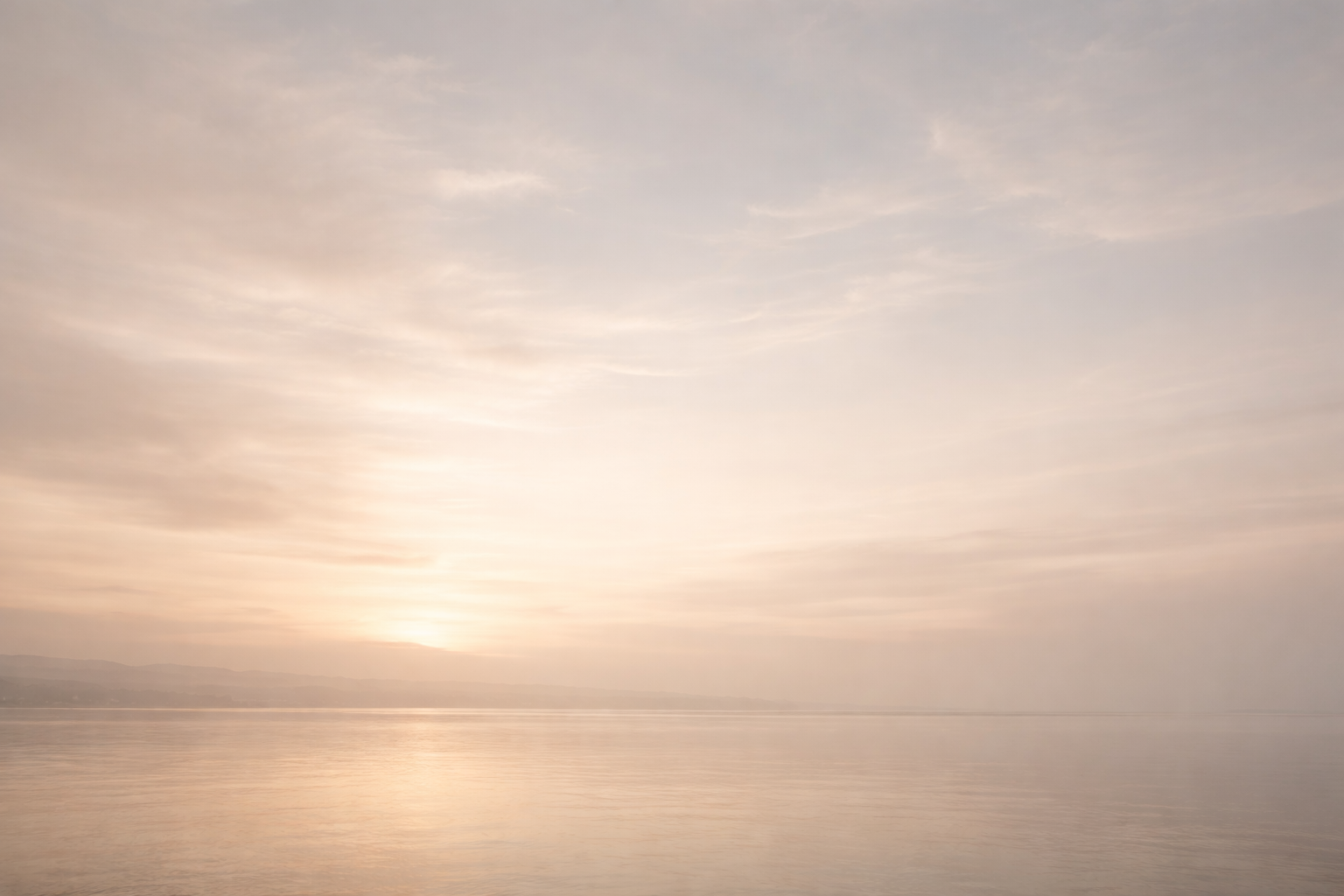Sunset over calm water with soft clouds and distant mountainous horizon.