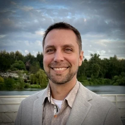 Smiling man in a light gray blazer standing outdoors with trees, a river, and cloudy sky in the background