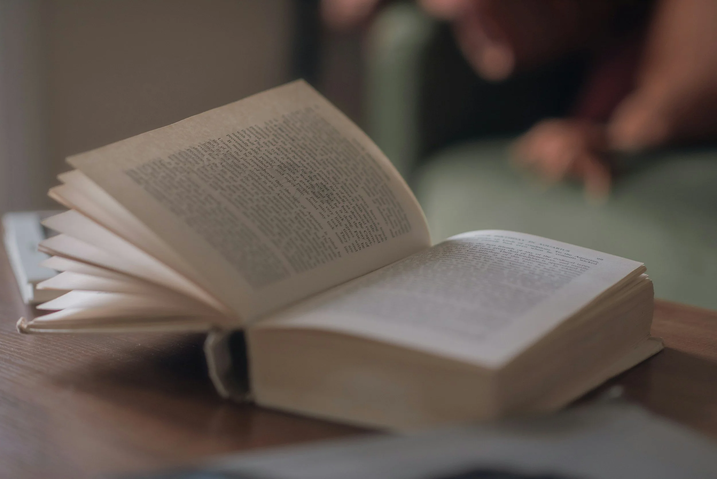 An open book resting on a wooden table, with a person in the background looking at it, out of focus.