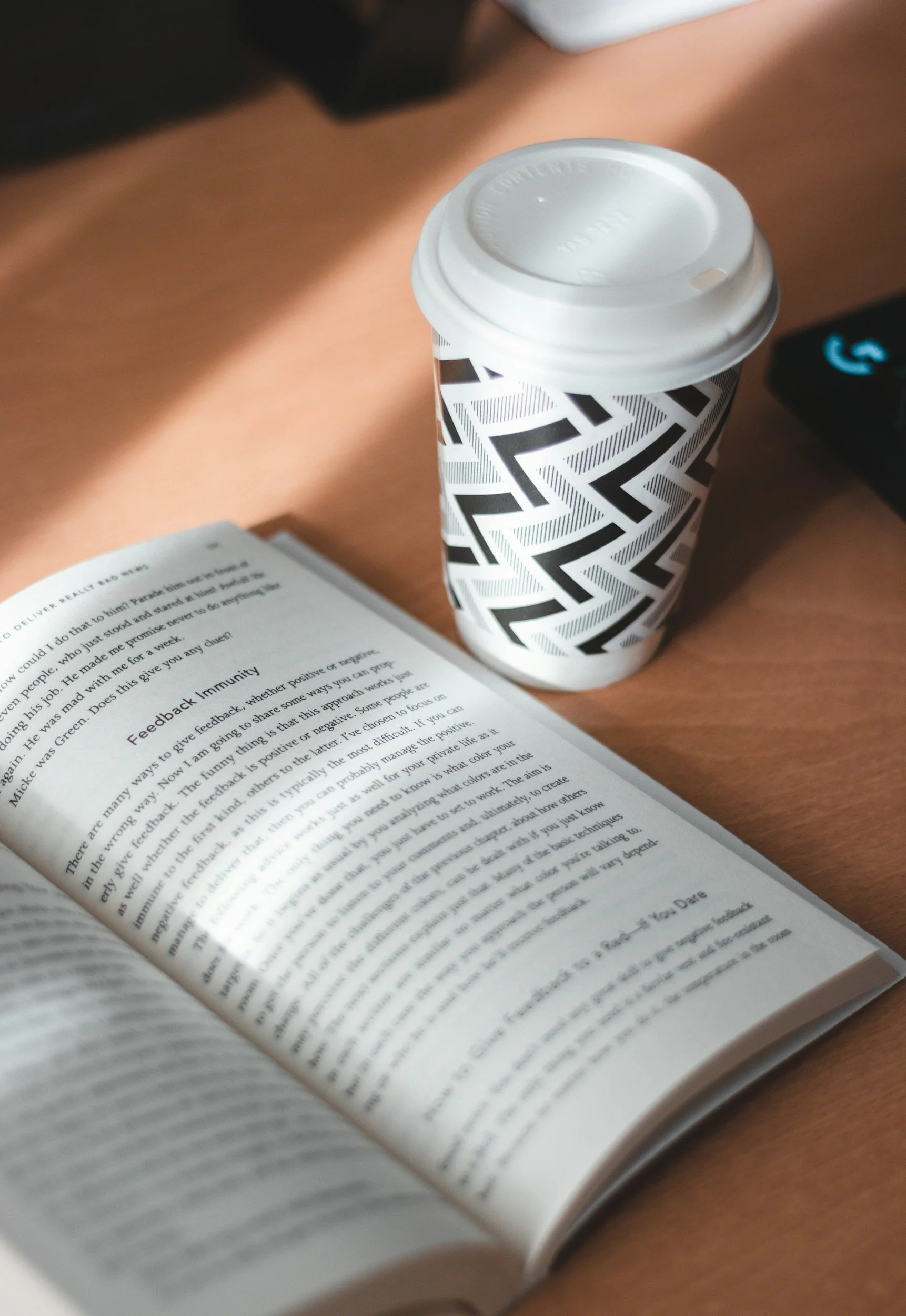 A paper coffee cup with a black and white geometric pattern, placed on a wooden table next to an open book with small text.