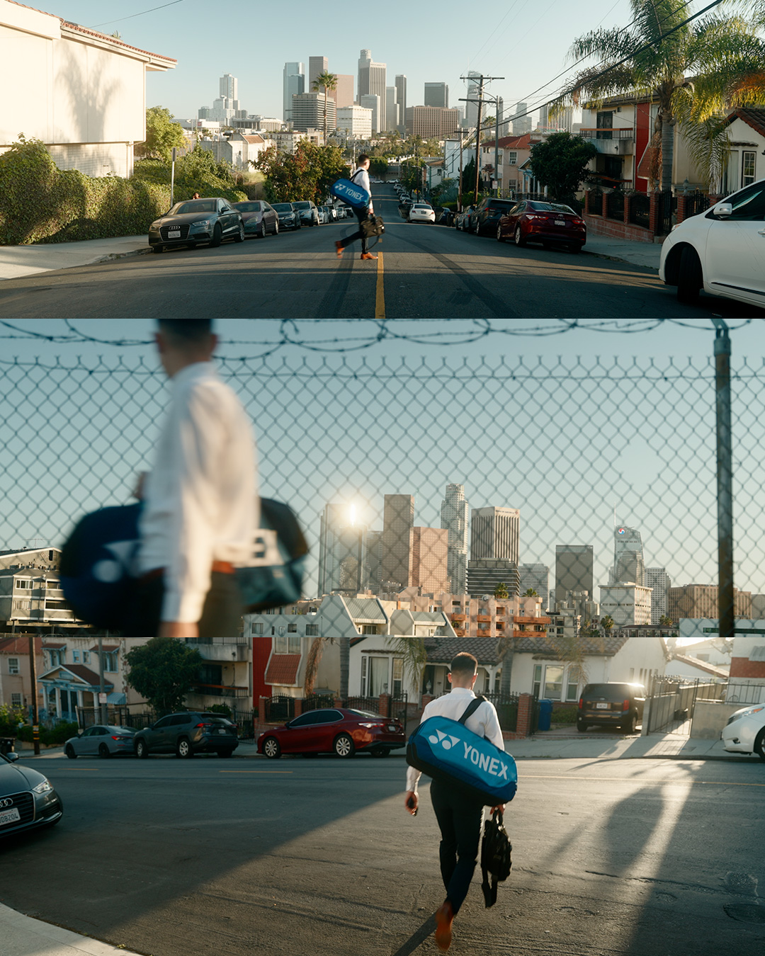 A man walking away carrying a blue YONEX sports bag and a black bag on a city street, with a view of downtown skyscrapers in the background, and a chain-link fence with another person in front of it.