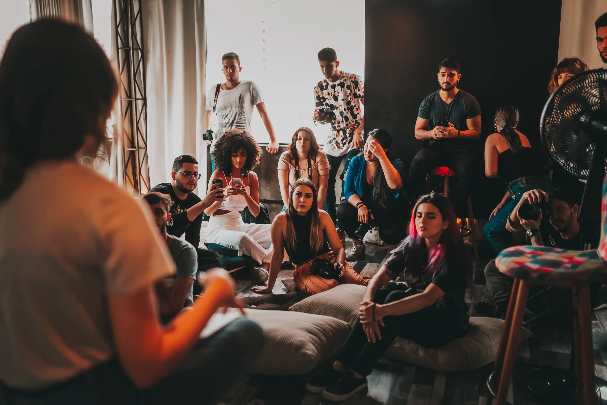 Group of diverse young people sitting and standing in a room, some using smartphones, with a person speaking in the foreground, and a large fan on the right side.