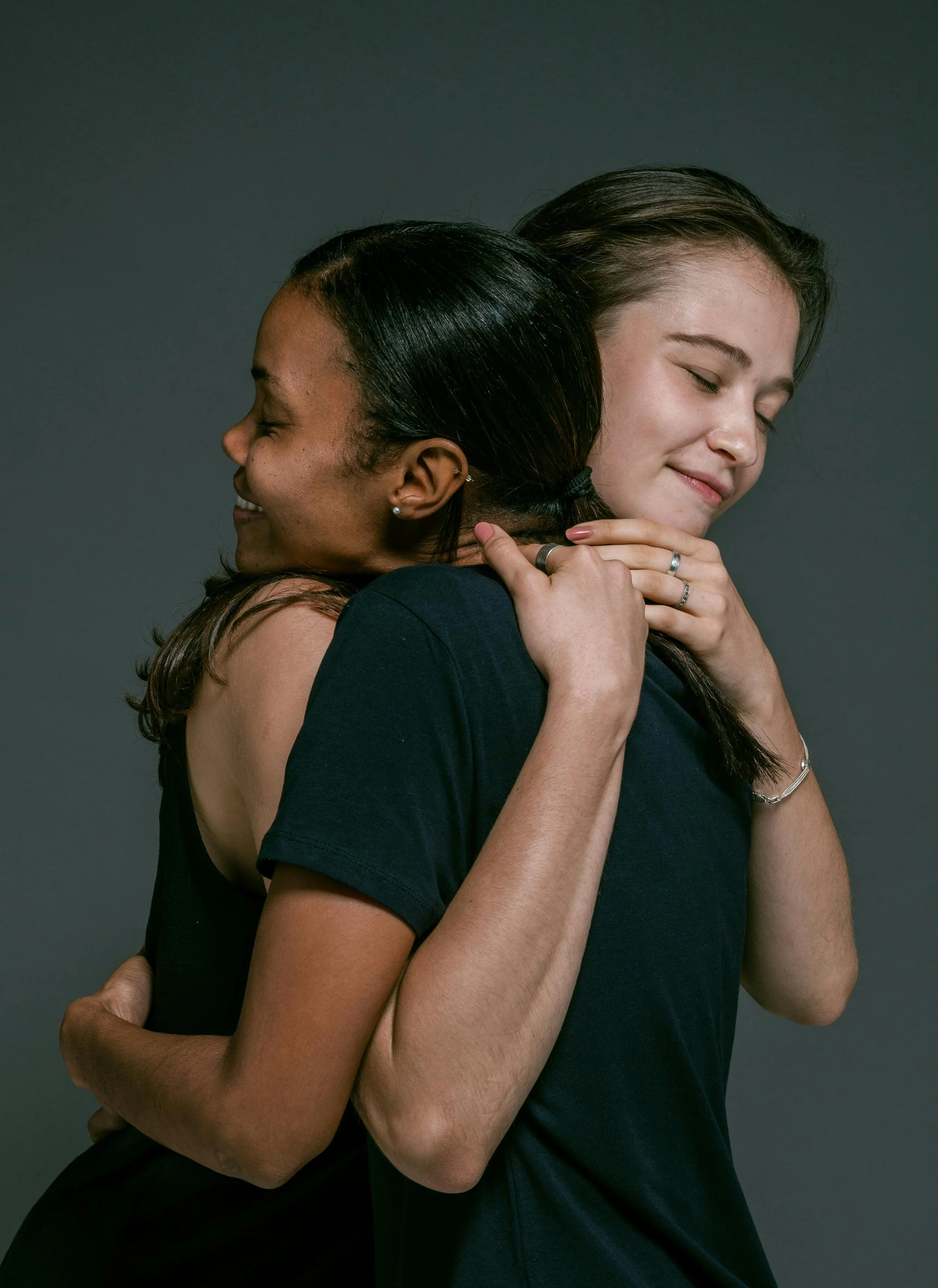 Two women hugging each other with closed eyes and smiles, against a dark background.