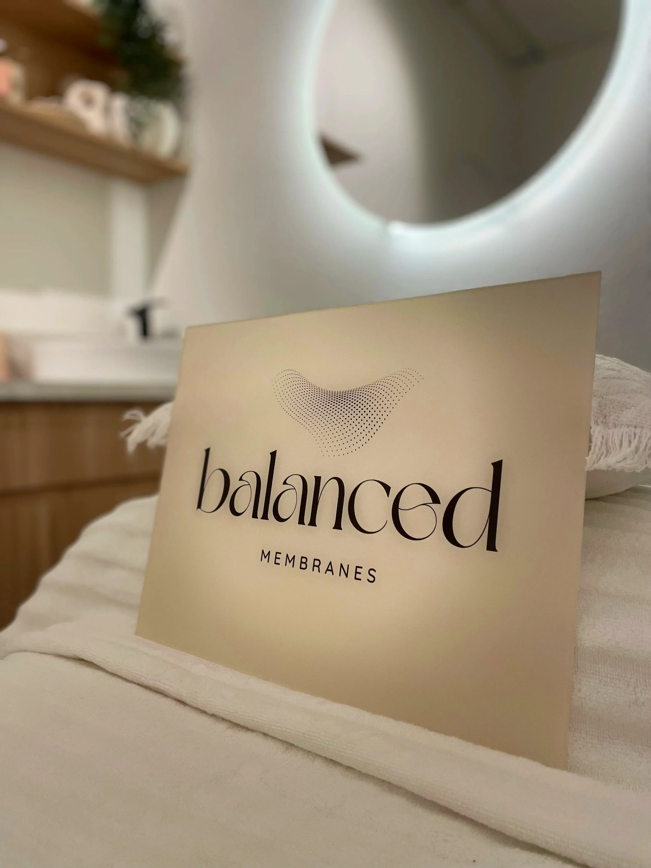 A sign reading 'balanced membranes' placed on a white surface in a room with a mirror and shelves in the background.