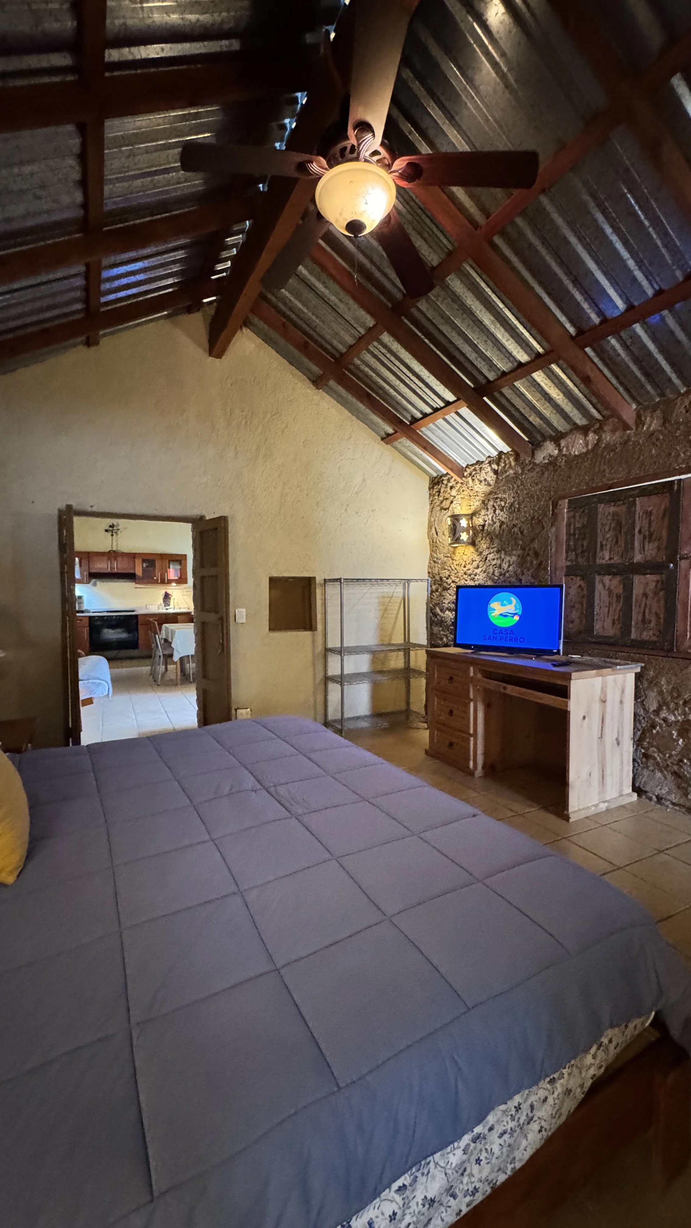 Interior de una habitación con techo de madera y lamina de metal, cama con sábanas grises, televisor sobre mueble de madera, paredes de piedra y algunos muebles de metal, vista hacia la cocina y comedor al fondo.