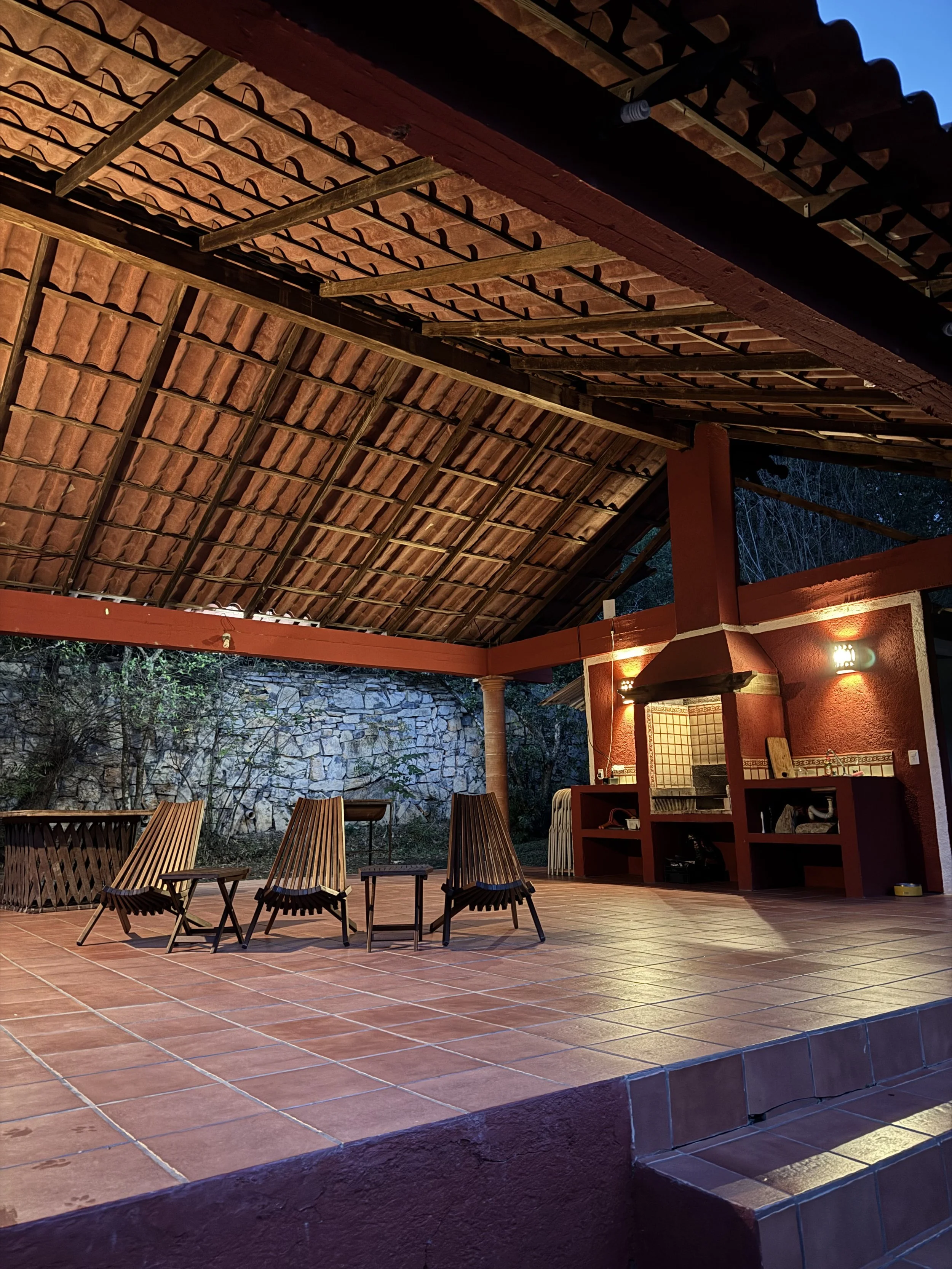 Un espacio al aire libre con piso de cerámica terracota, paredes de piedra y techo de tejas de barro, con mobiliario de madera y un área de cocina con chimenea, iluminado por luces cálidas en la noche.
