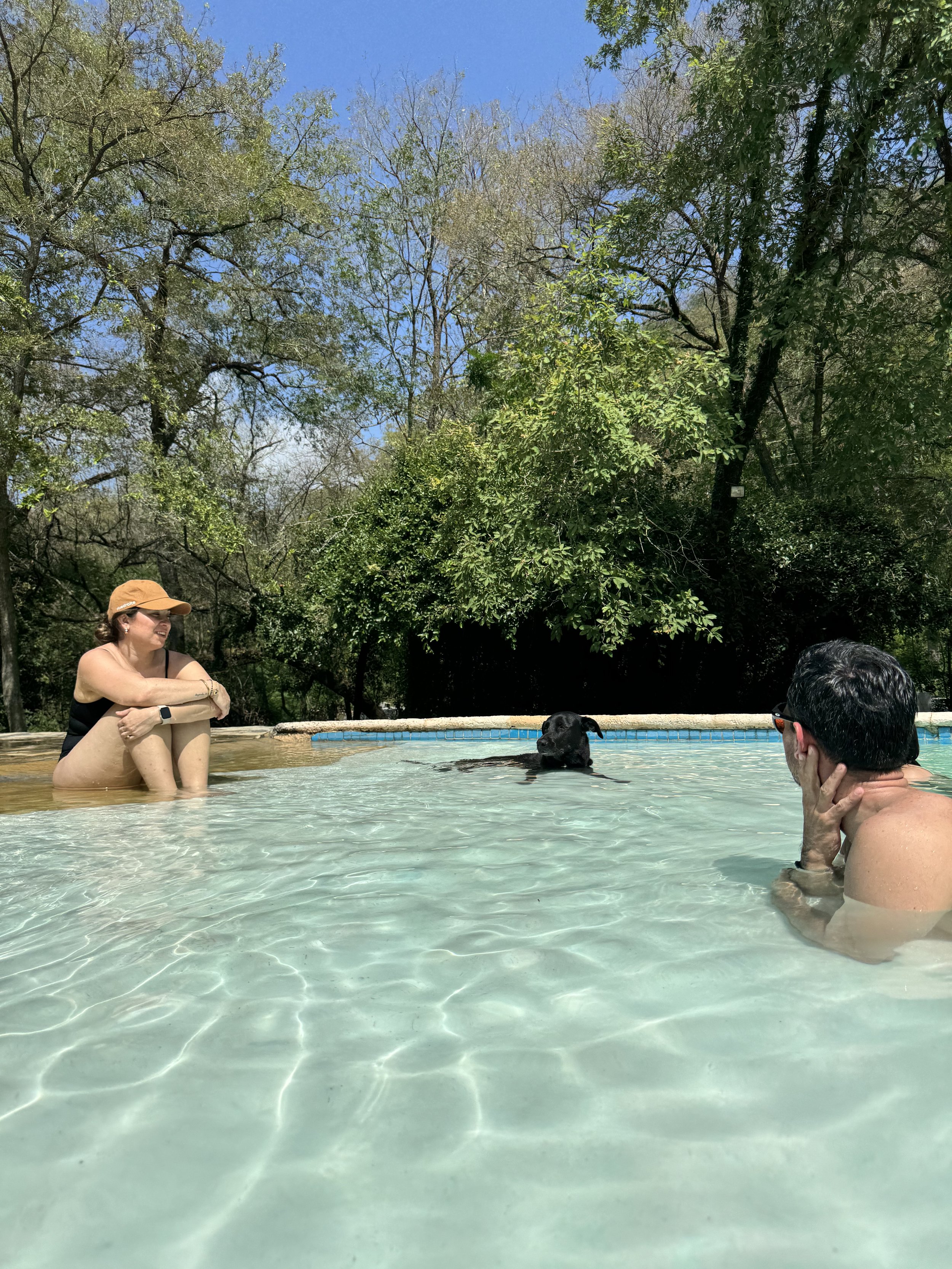 Dos personas y un perro en una piscina al aire libre, rodeados de árboles y cielo azul.