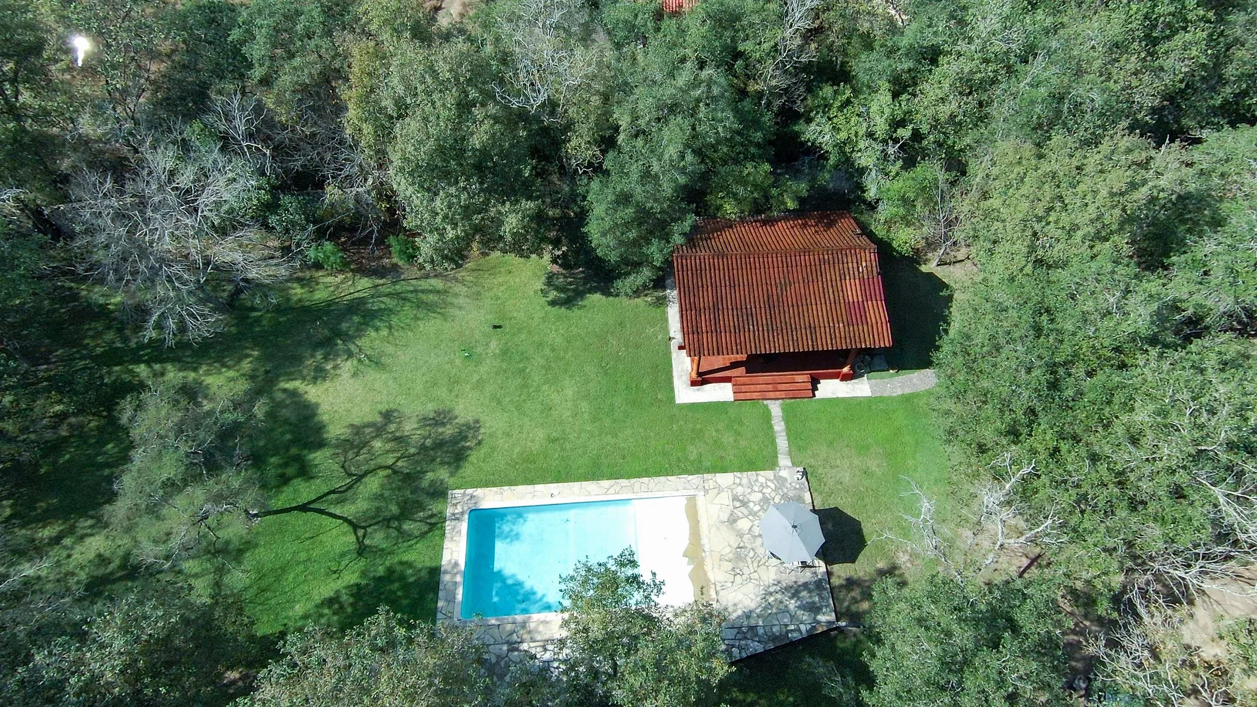 Vista aérea de un área con un césped verde, un pequeño edificio con techo de tejas rojas y una piscina de agua azul rodeada de piedras, rodeado de árboles.