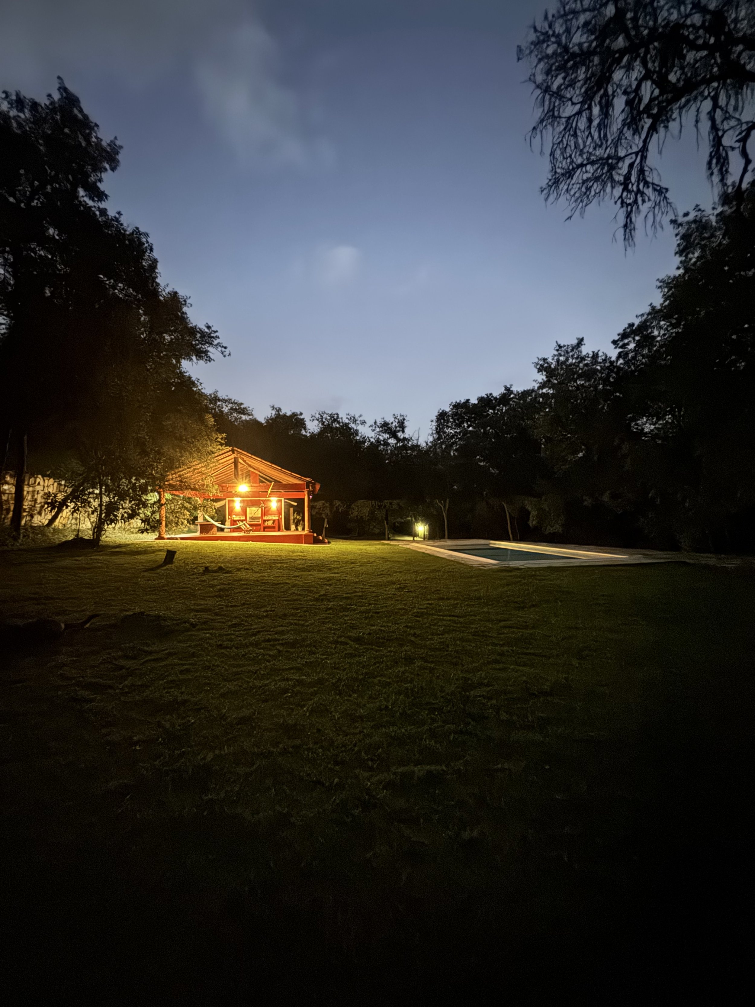 Escena nocturna en un jardín con césped, árboles y una piscina. Hay una pequeña estructura de madera con iluminación cálida, y en el fondo se ve el cielo con algunas nubes