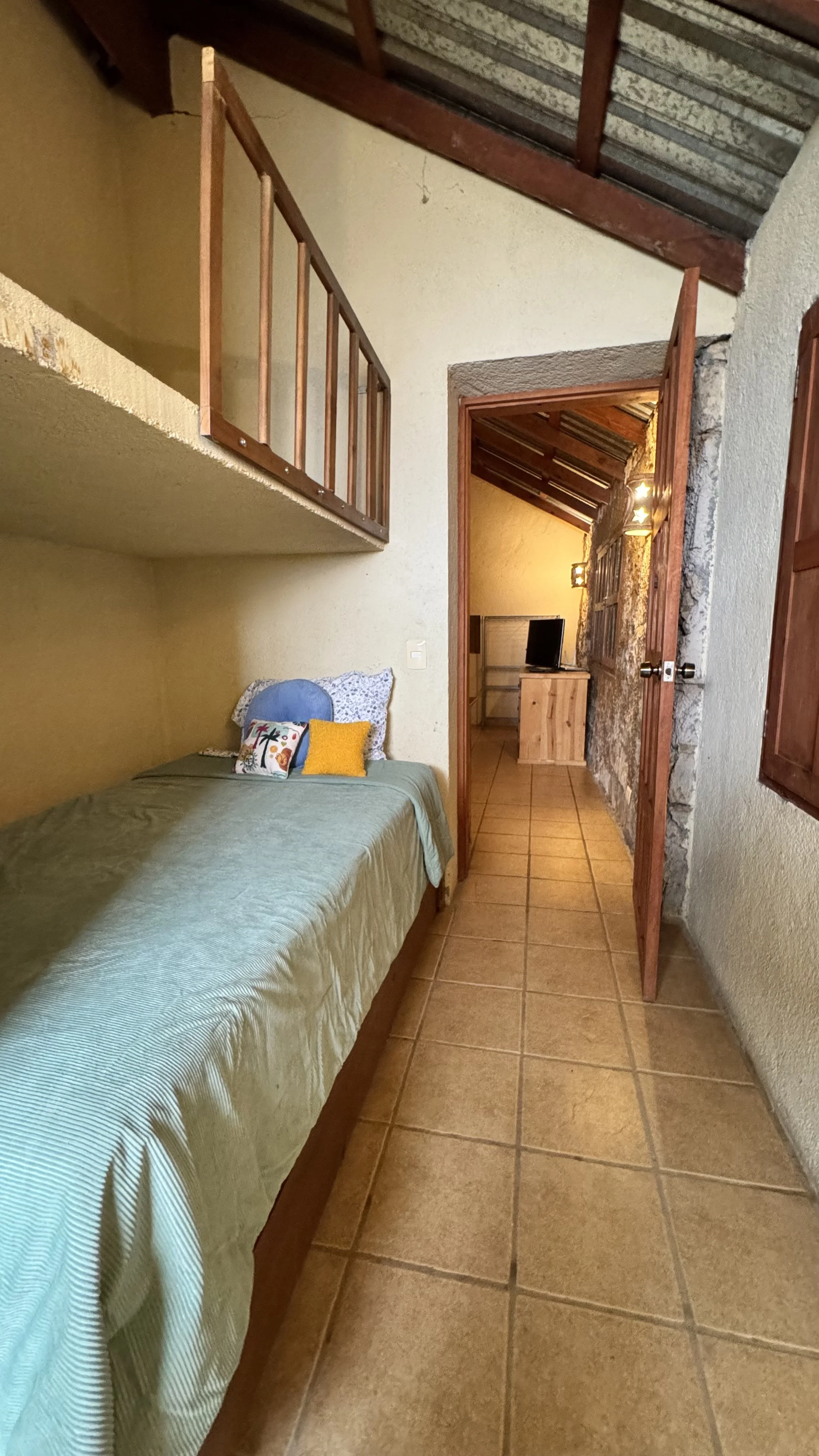 Dormitorio con cama, cojines, paredes de colores cálidos, suelo de baldosas, puerta abierta a un pasillo con televisión y mobiliario de madera.