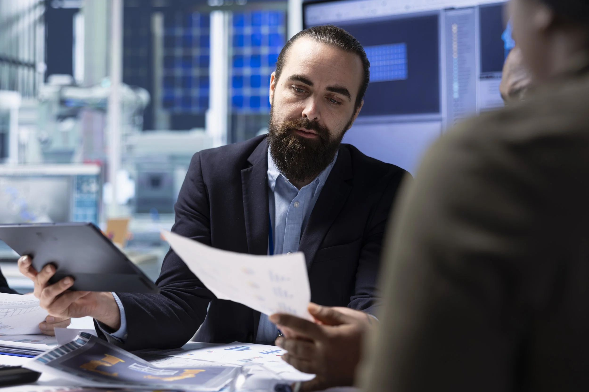 Two men in business attire sit at a table reviewing documents in a modern office with large screens and graphs in the background.