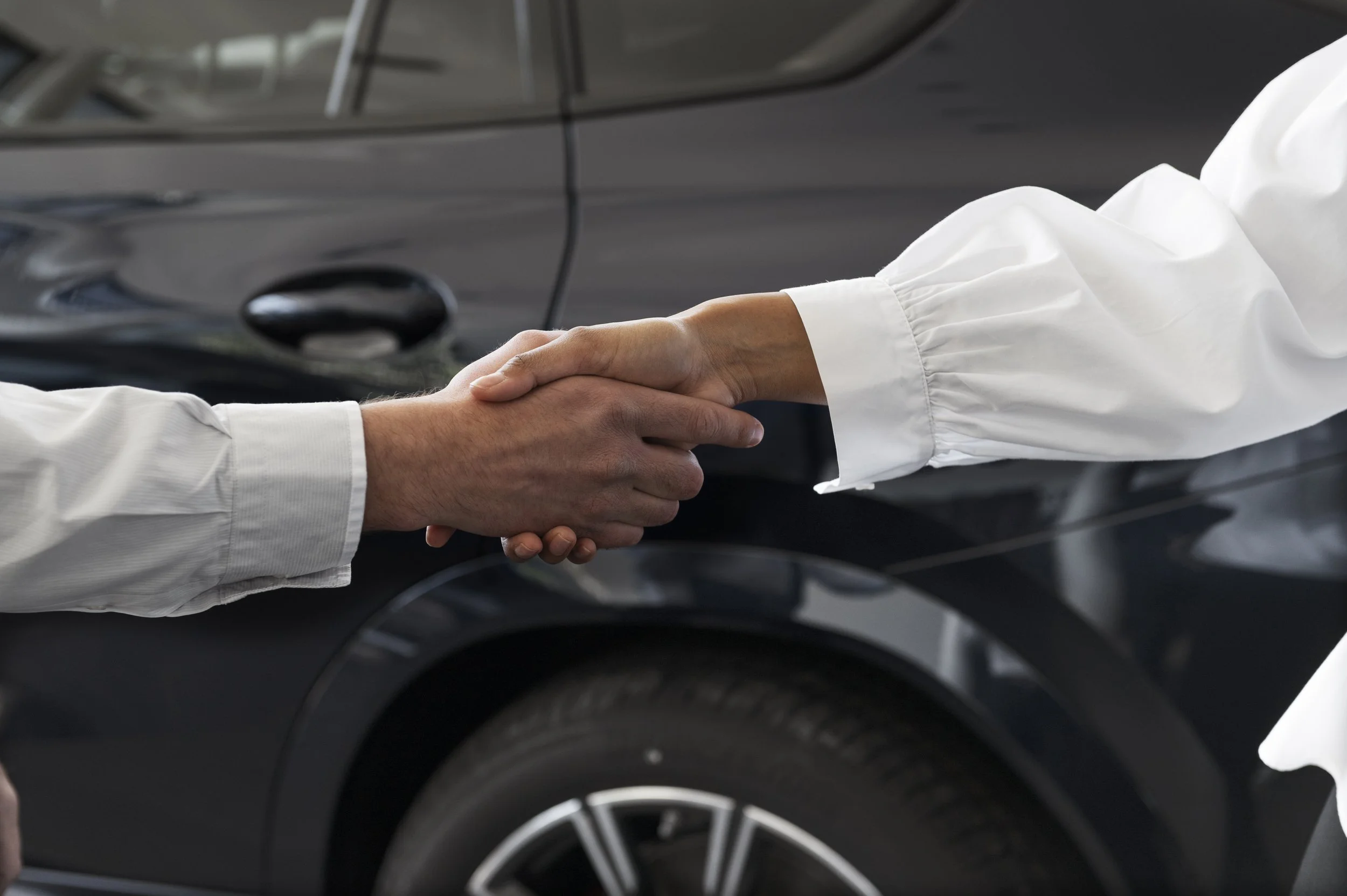 Two people shaking hands in front of a black car outside.