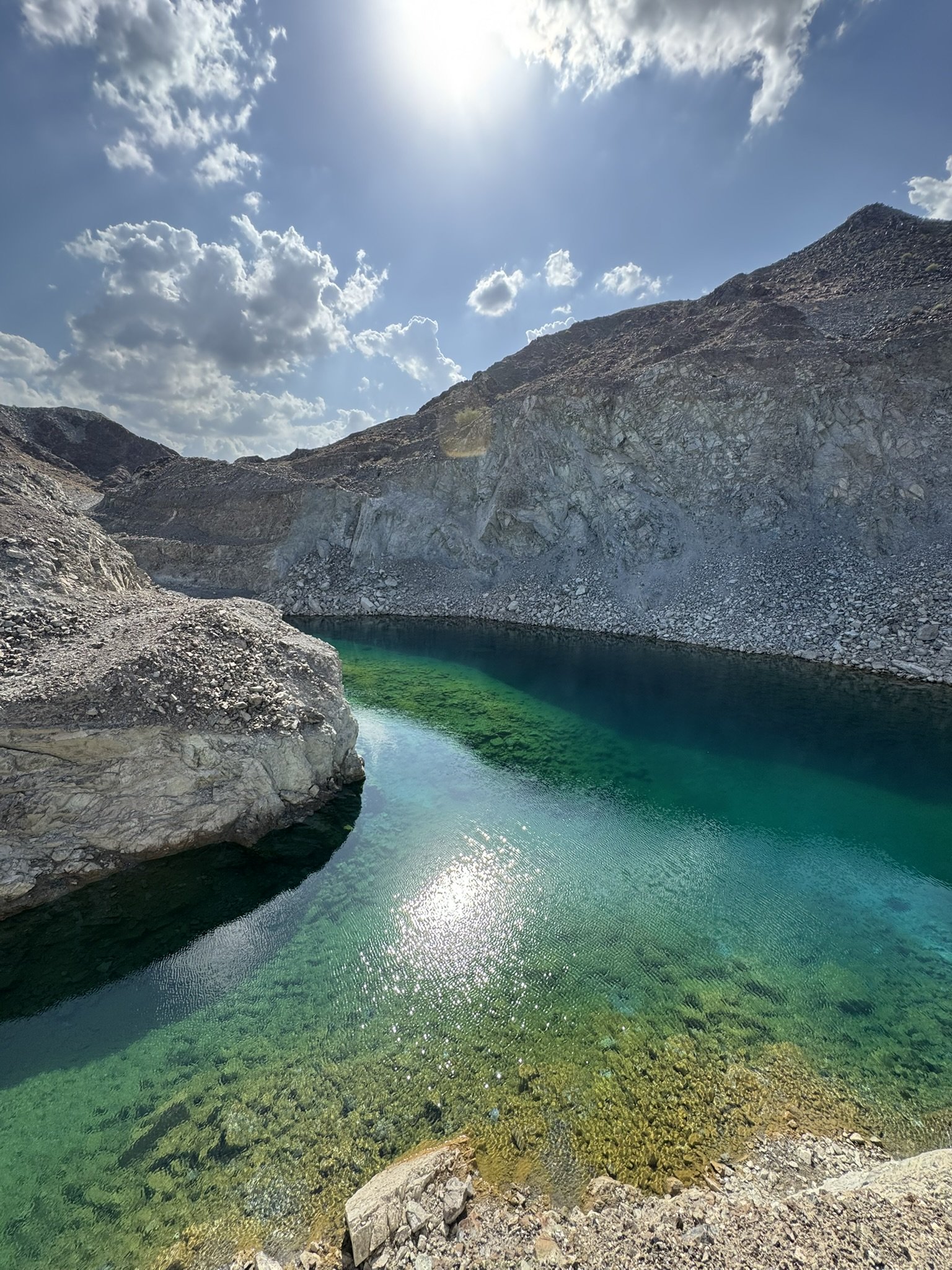 A serene mountain lake with clear green water, surrounded by rocky slopes under a partly cloudy sky with the sun shining brightly.