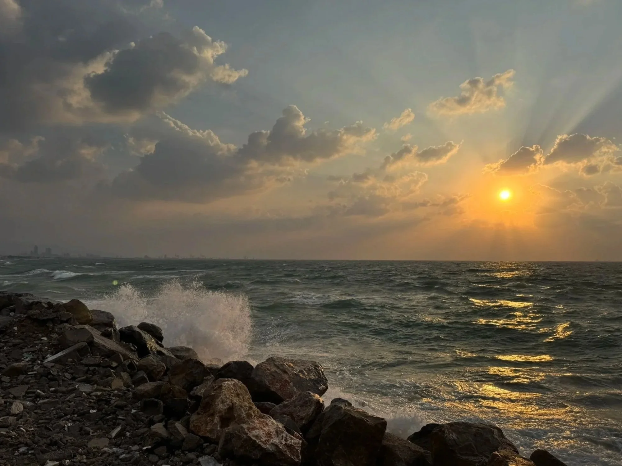 Sun setting over the ocean with waves crashing against rocks on the shoreline and clouds in the sky.