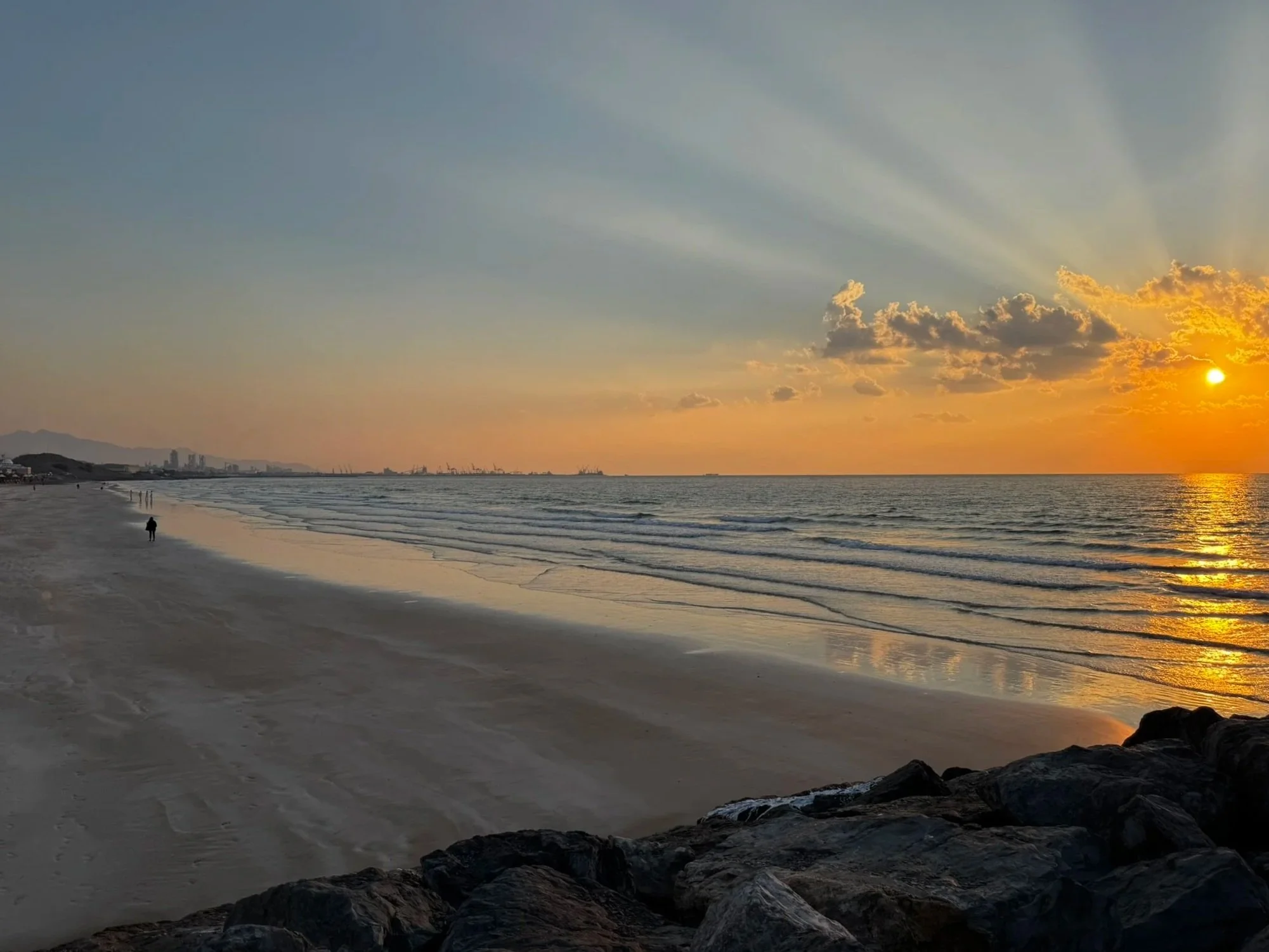 Sunset over a calm beach with a few people walking along the shoreline, rocks in the foreground, and cityscape with cranes in the distance.