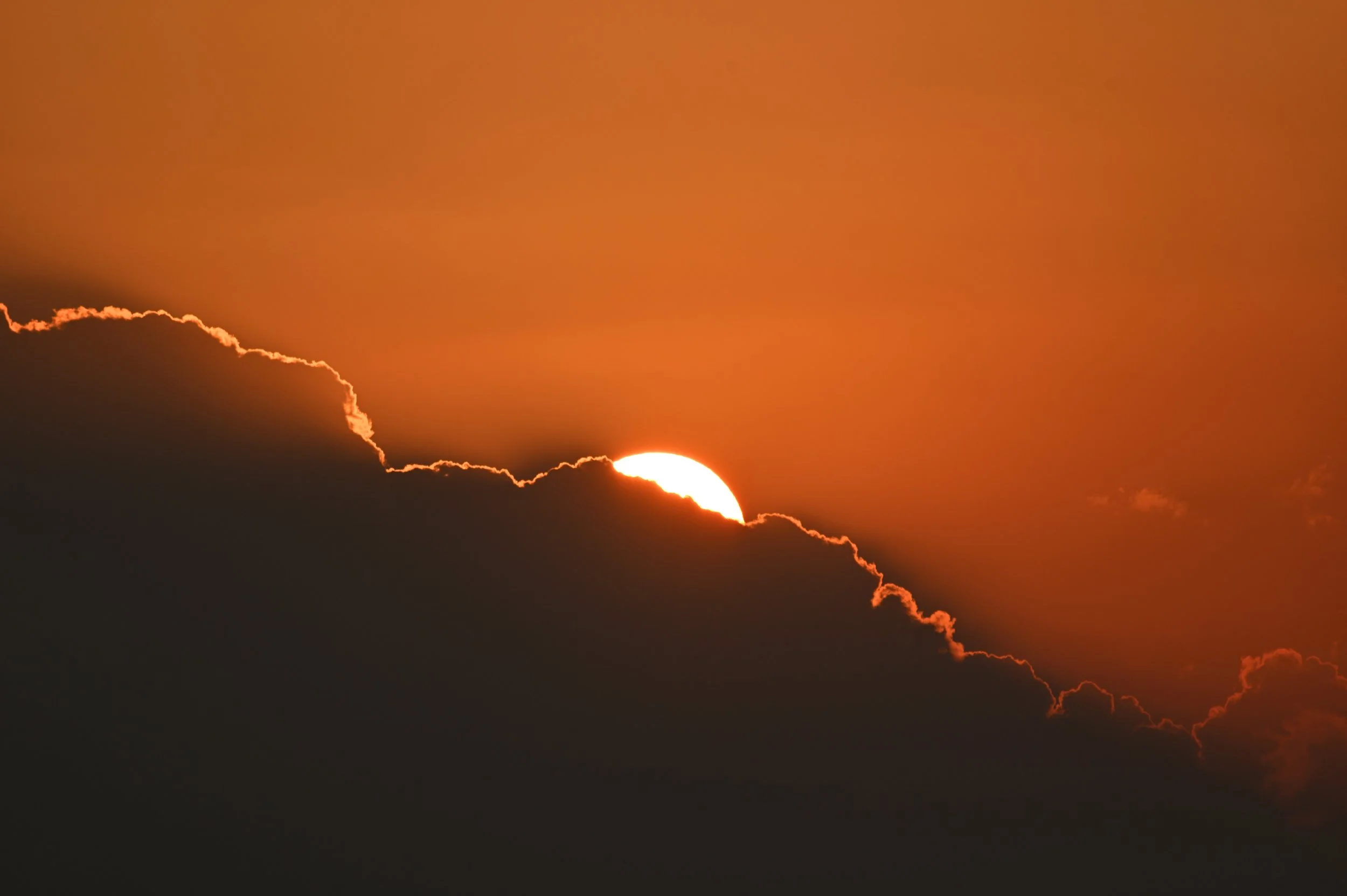 Sunset partially obscured by dark clouds, creating a glowing edge around the clouds with a warm orange sky.