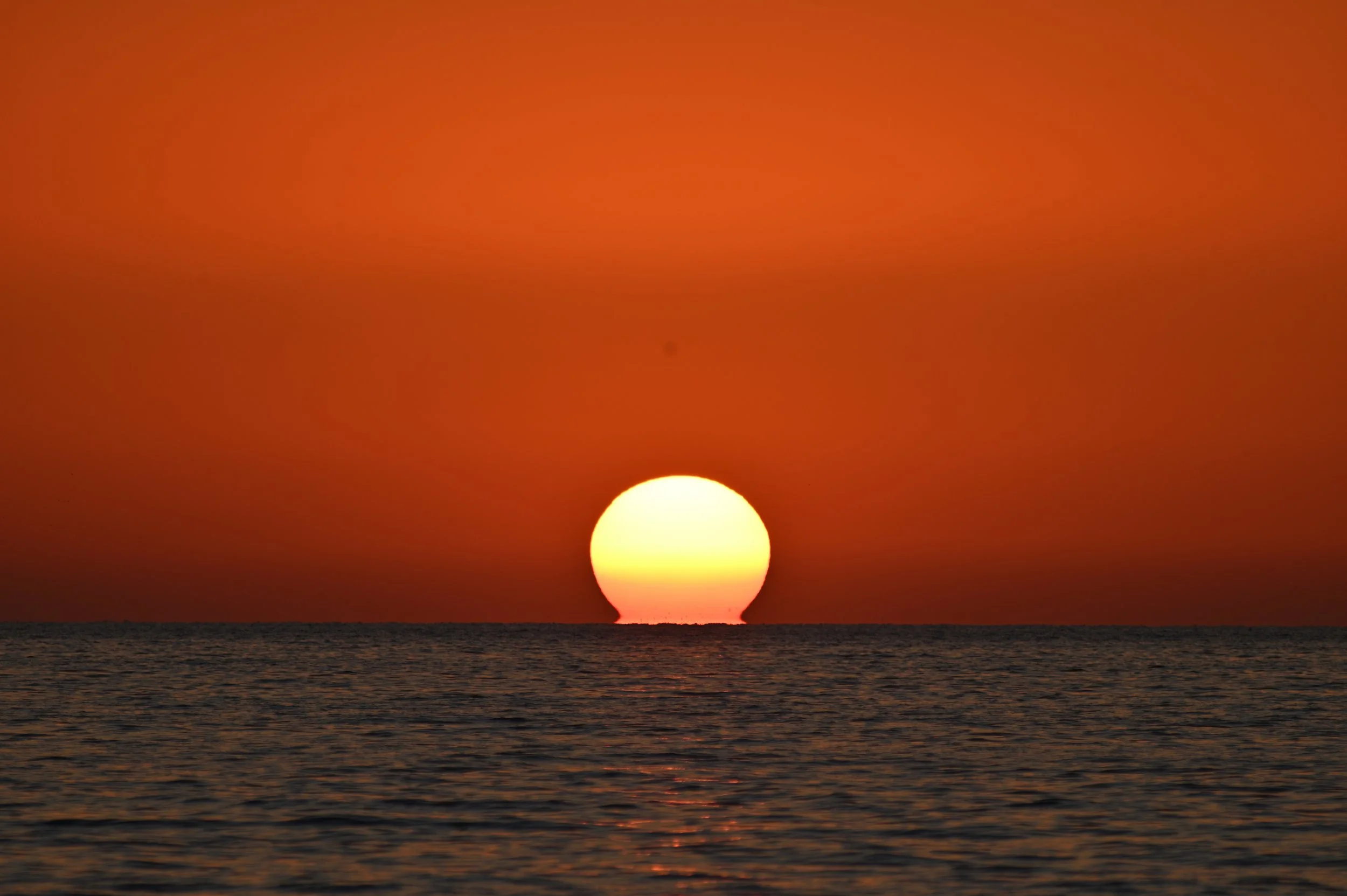 Sunset over the ocean with the sun partially below the horizon, casting an orange glow in the sky.