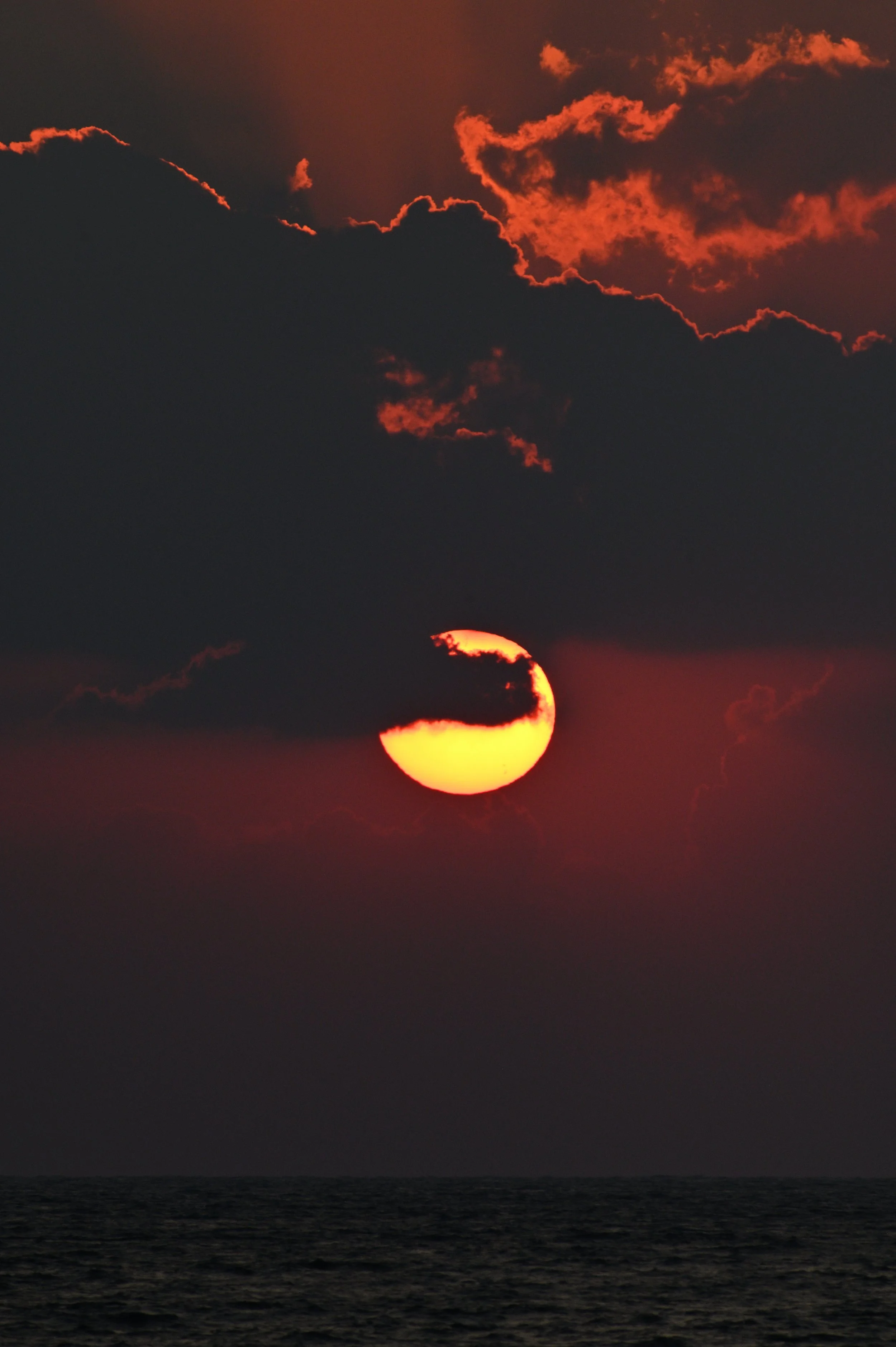 Sunset over the ocean with dark clouds partially covering the sun and a reddish-orange sky.
