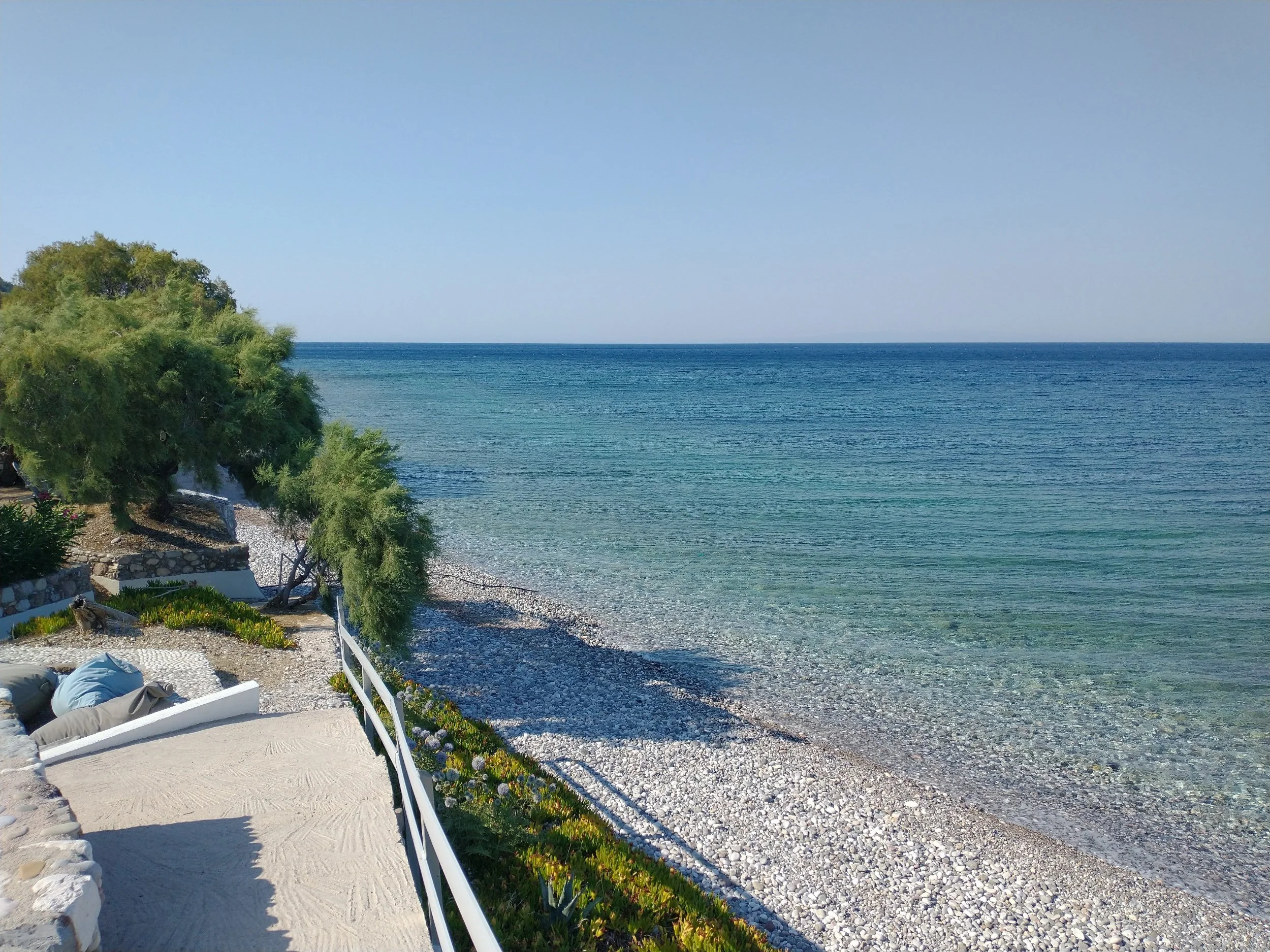 Scenic view of a pebbled beach with clear blue water and a green tree on the left side under a clear sky.