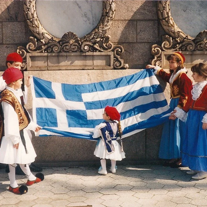 Children dressed in traditional Greek costumes stand outdoors holding a Greek flag.