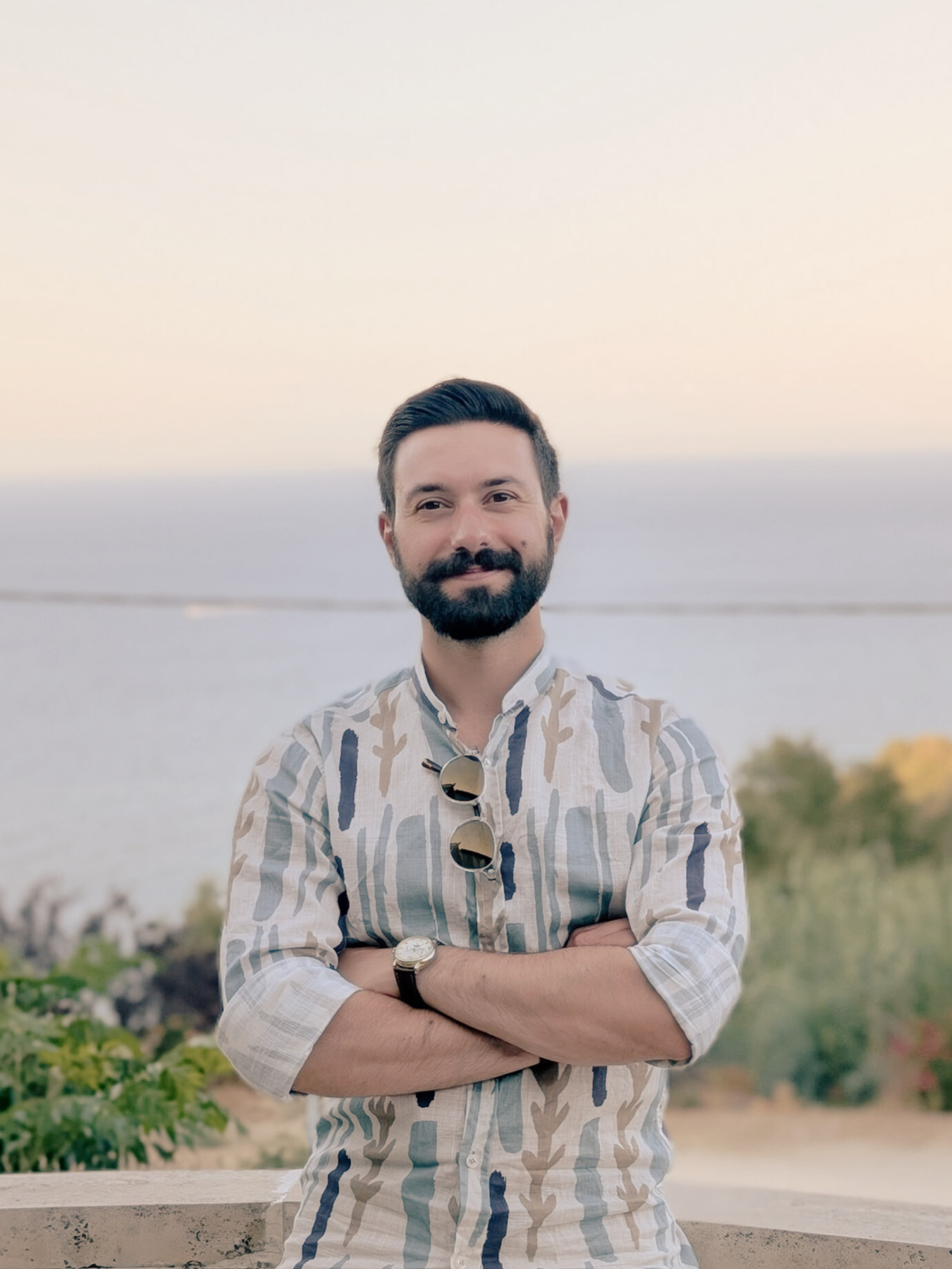 A man with a beard and mustache standing outdoors near a body of water at sunset, wearing a patterned shirt with sunglasses hanging on his collar and a wristwatch, with his arms crossed.