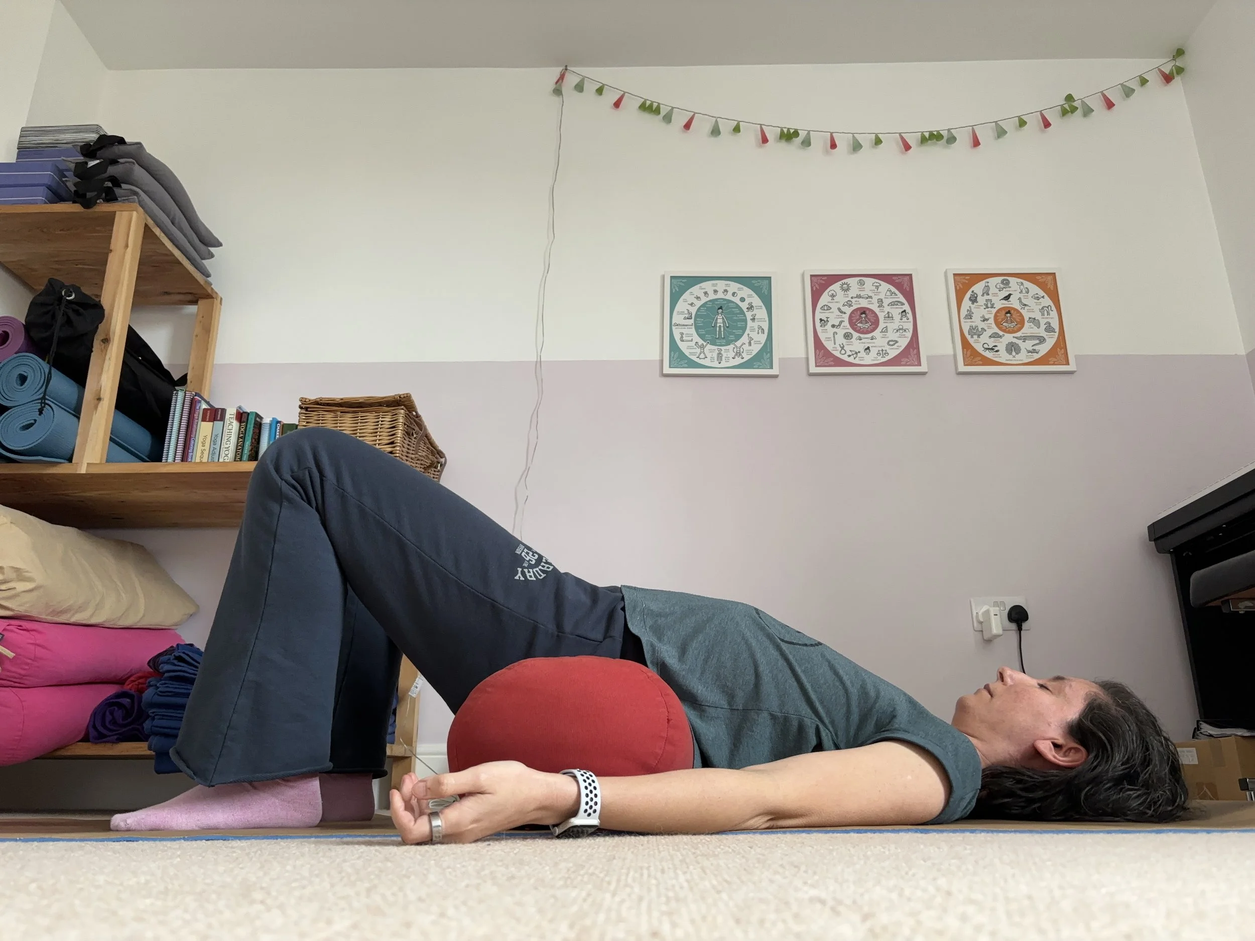 A person lying on their back on a yoga mat in a room, with knees bent and supported by a red bolster, practicing yoga or relaxation.
