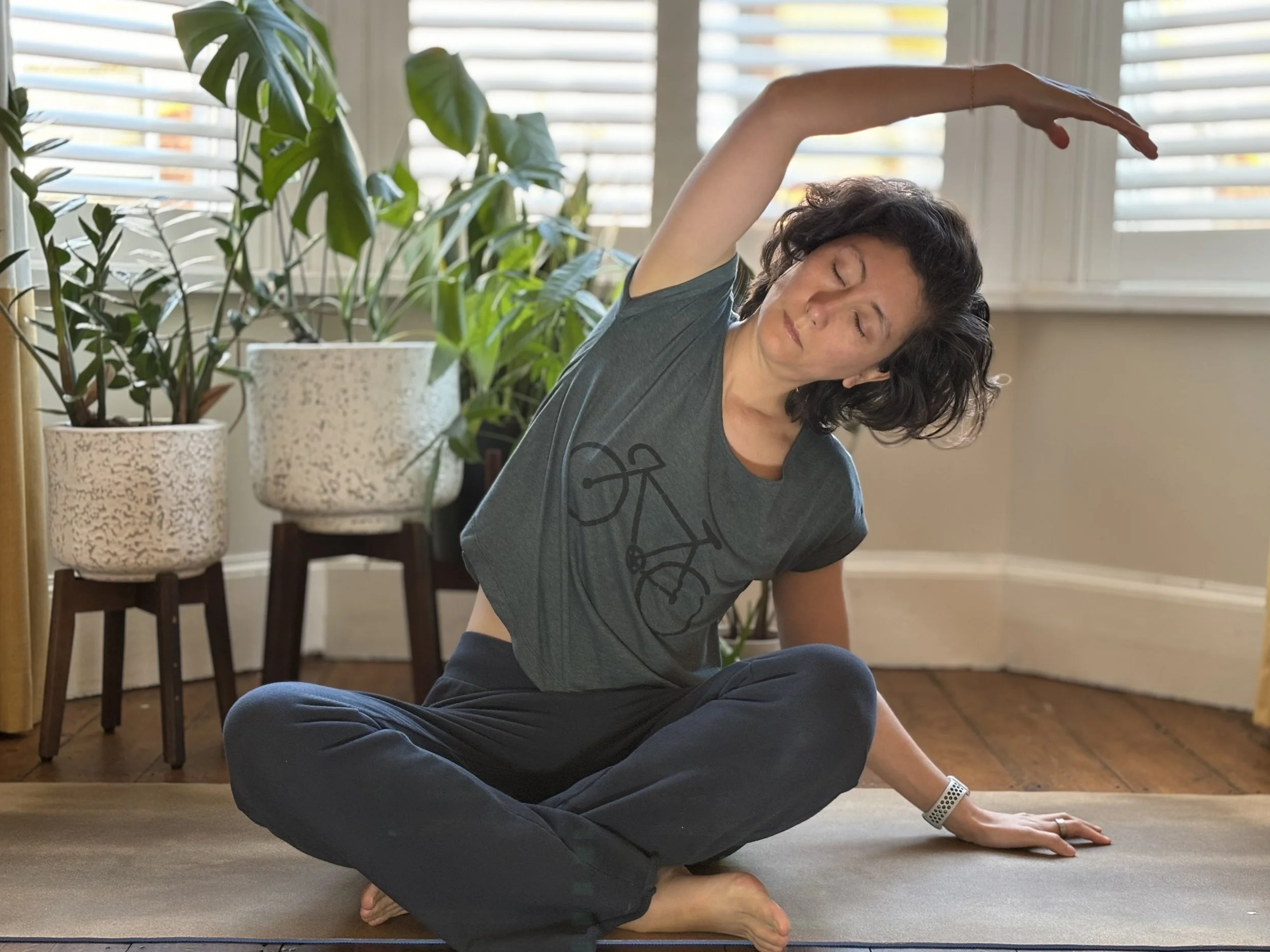 A woman practicing yoga indoors near large potted plants, sitting cross-legged on a mat, stretching her left arm over her head.