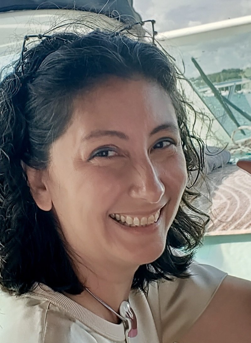 A smiling woman with curly dark hair, sitting on a boat with water and another boat visible in the background.