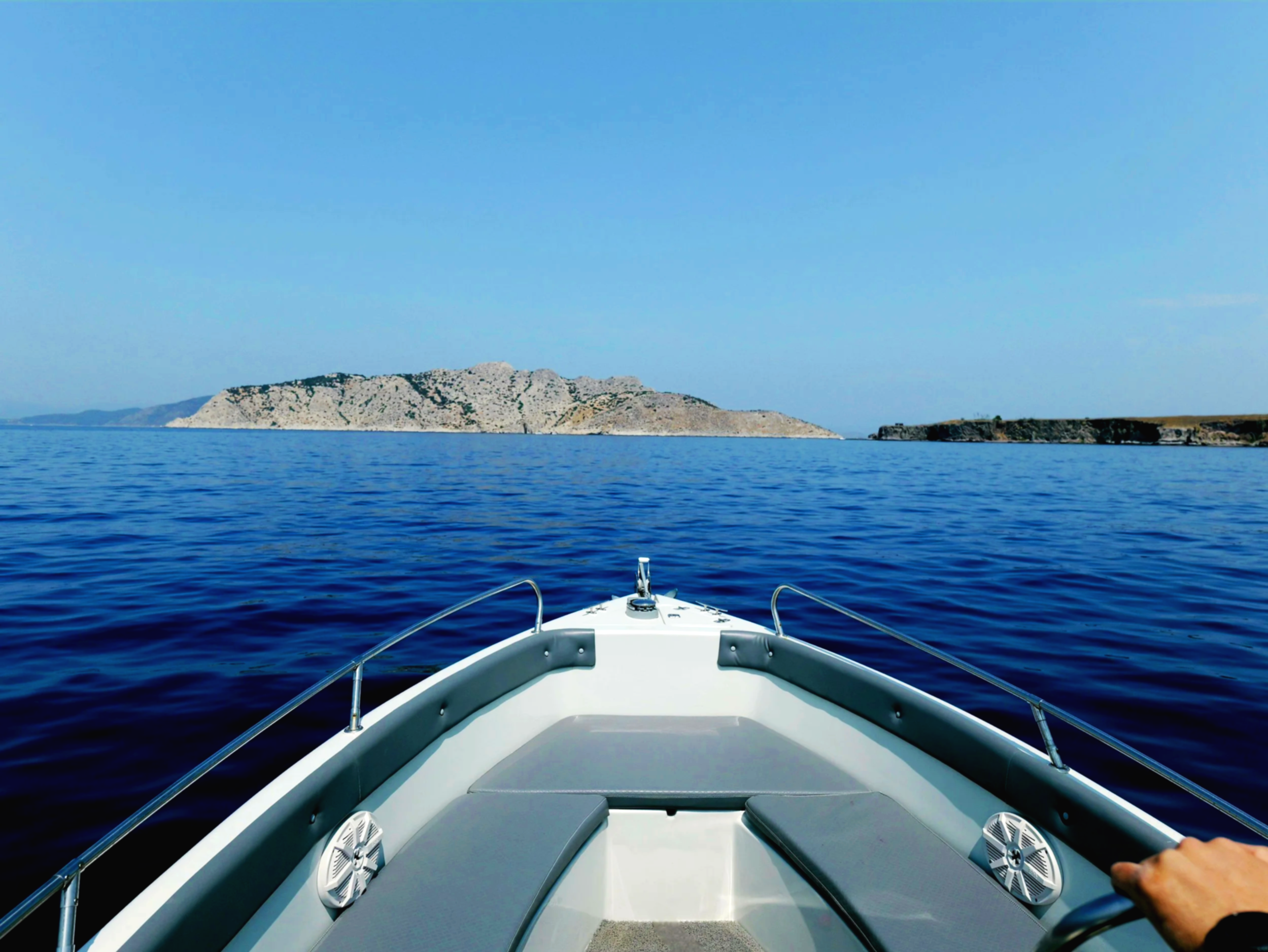 View from the front of a boat cruising on calm blue ocean waters towards distant rocky islands and coastline under a clear blue sky.