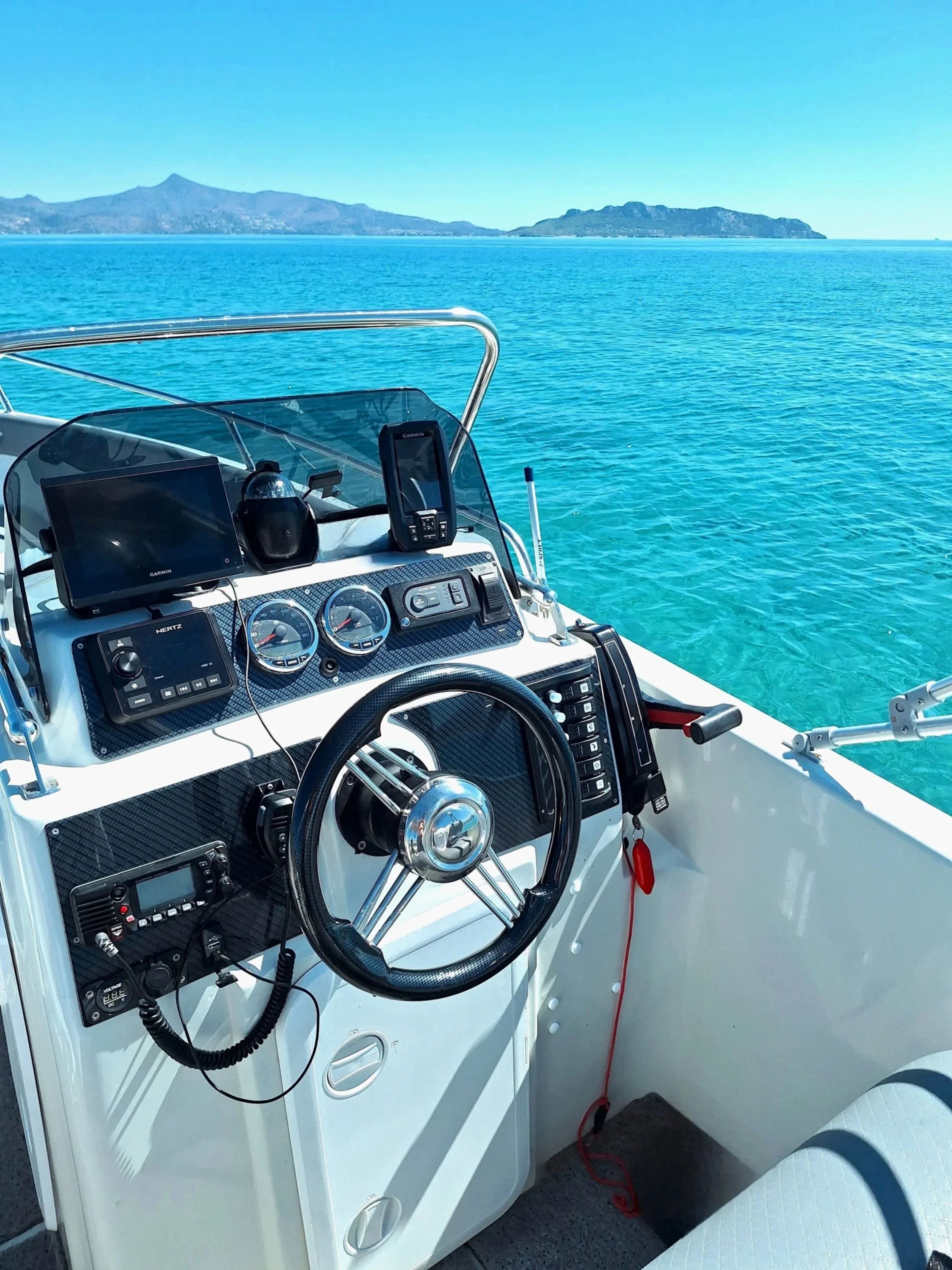 View of a boat's dashboard with various gauges, GPS, and communication devices, overlooking clear turquoise water and distant mountains.