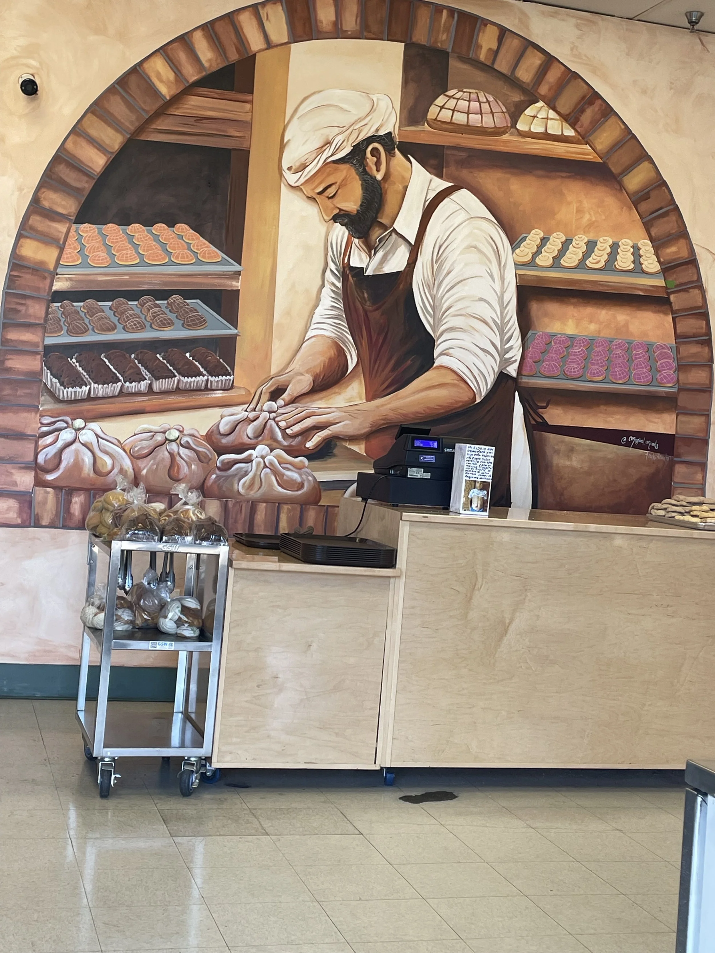 A mural of a man with a beard and turban making pastries behind a bakery counter. The counter has packaged baked goods and a small cart with more baked items.