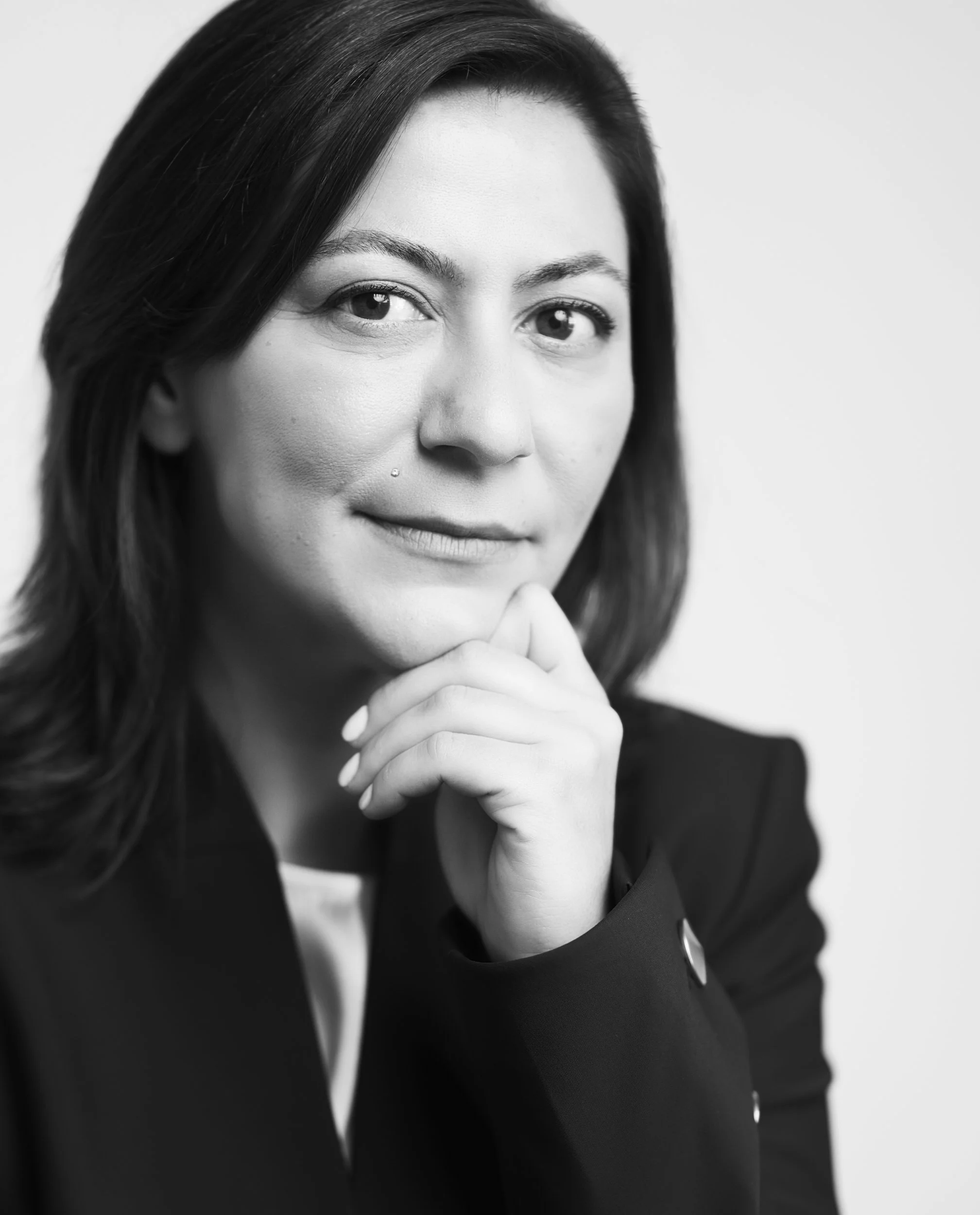 Black and white portrait of a woman with dark hair, resting her chin on her hand, wearing a dark blazer and a light-colored shirt, looking confidently at the camera.