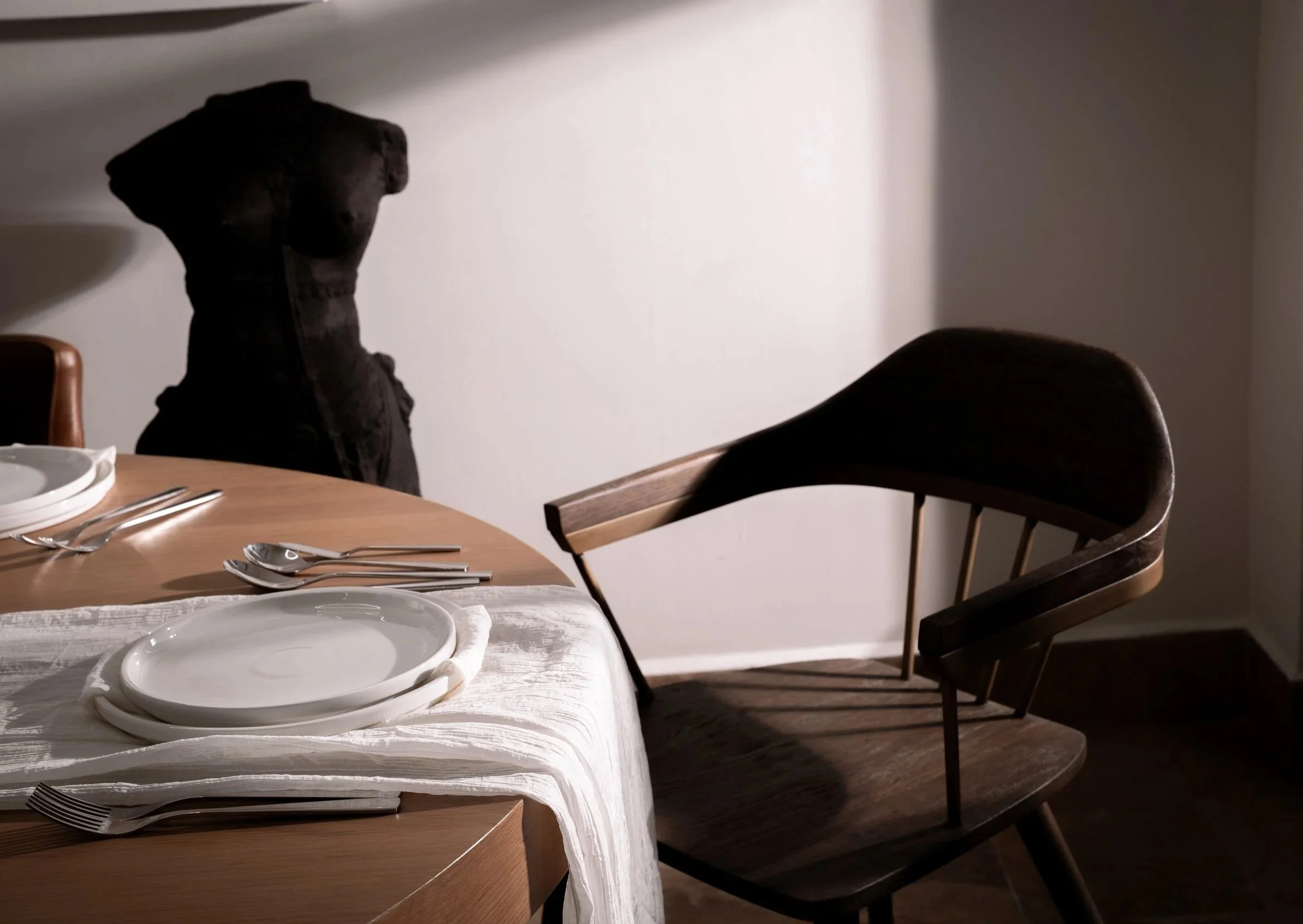 A dining table set with white plates, silverware, and a white tablecloth; a dark wooden chair is positioned next to the table, and a black jacket hangs on the back of a chair in the background.