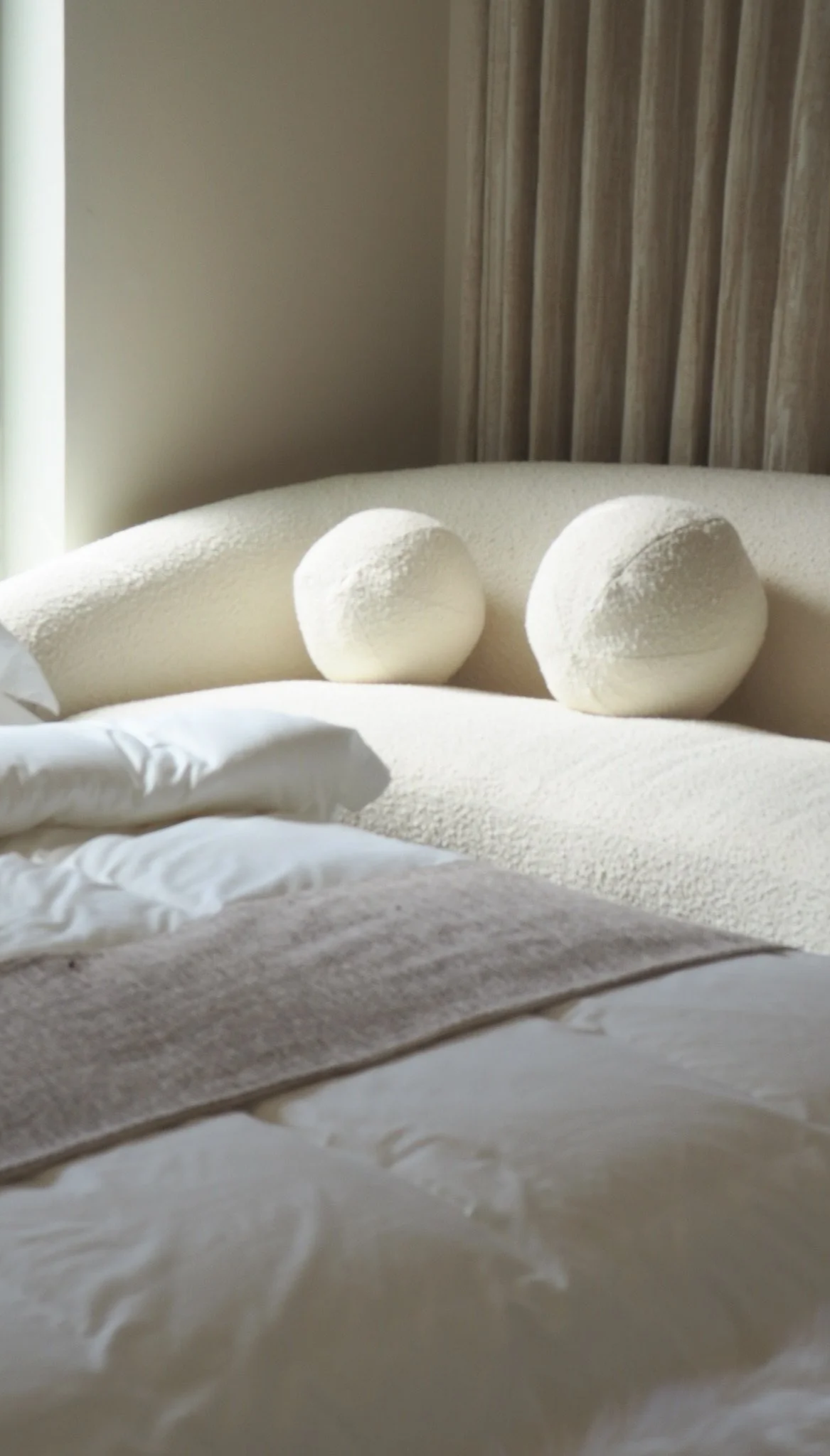 A bed with white sheets and pillows, and two white spherical decorative cushions on the headboard, in a softly lit bedroom