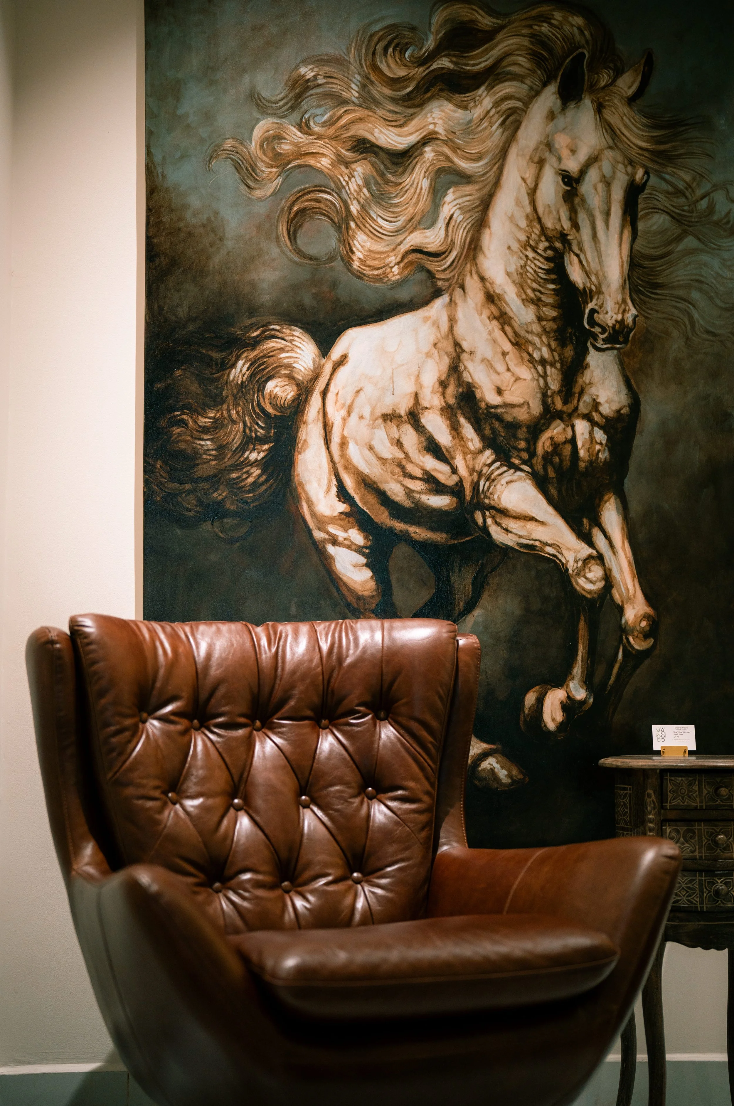 A large, detailed painting of a rearing horse with flowing mane on a dark background, located behind a brown leather armchair in a room.