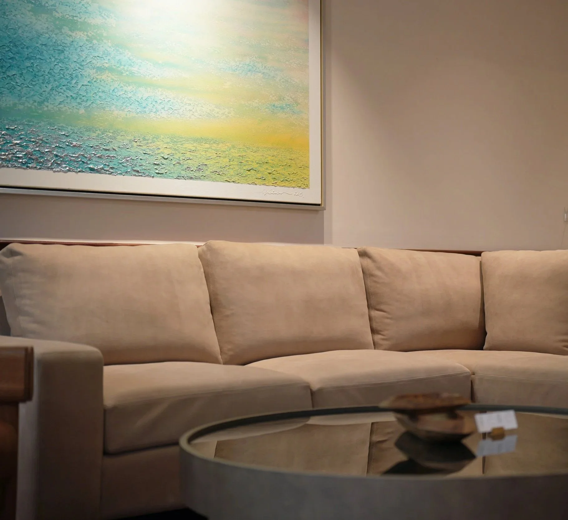 A beige sofa in a living room with a large, abstract landscape painting on the wall above it and a glass-top coffee table with a bowl and a card in front.