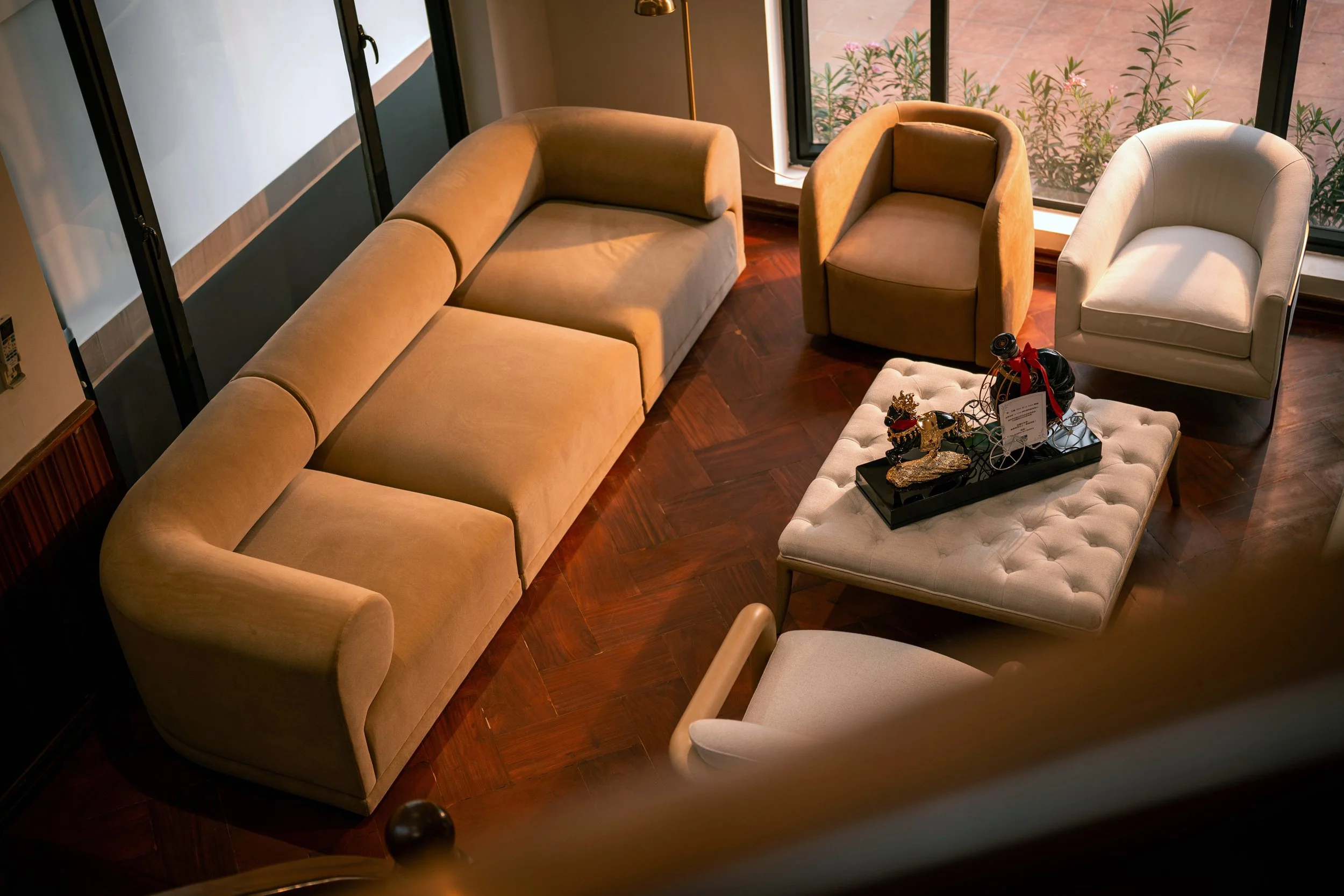 A cozy living room with a beige sofa, two accent chairs in tan and cream, a tufted ottoman with holiday decorations, and a large window showing outdoor plants.