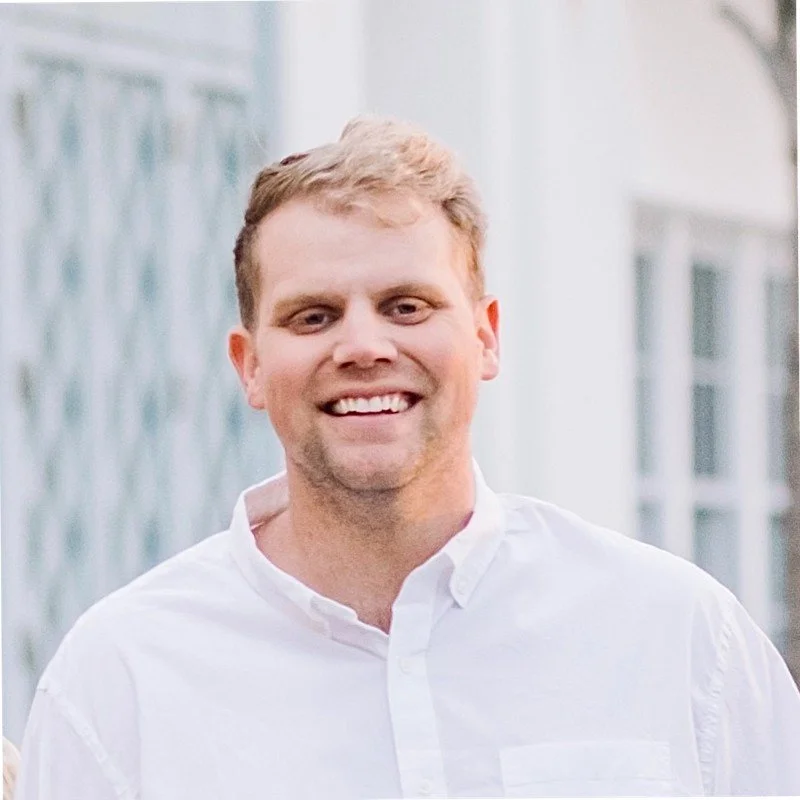 A man with light skin, short reddish-blonde hair, smiling, wearing a white button-up shirt, standing outdoors with a building in the background.