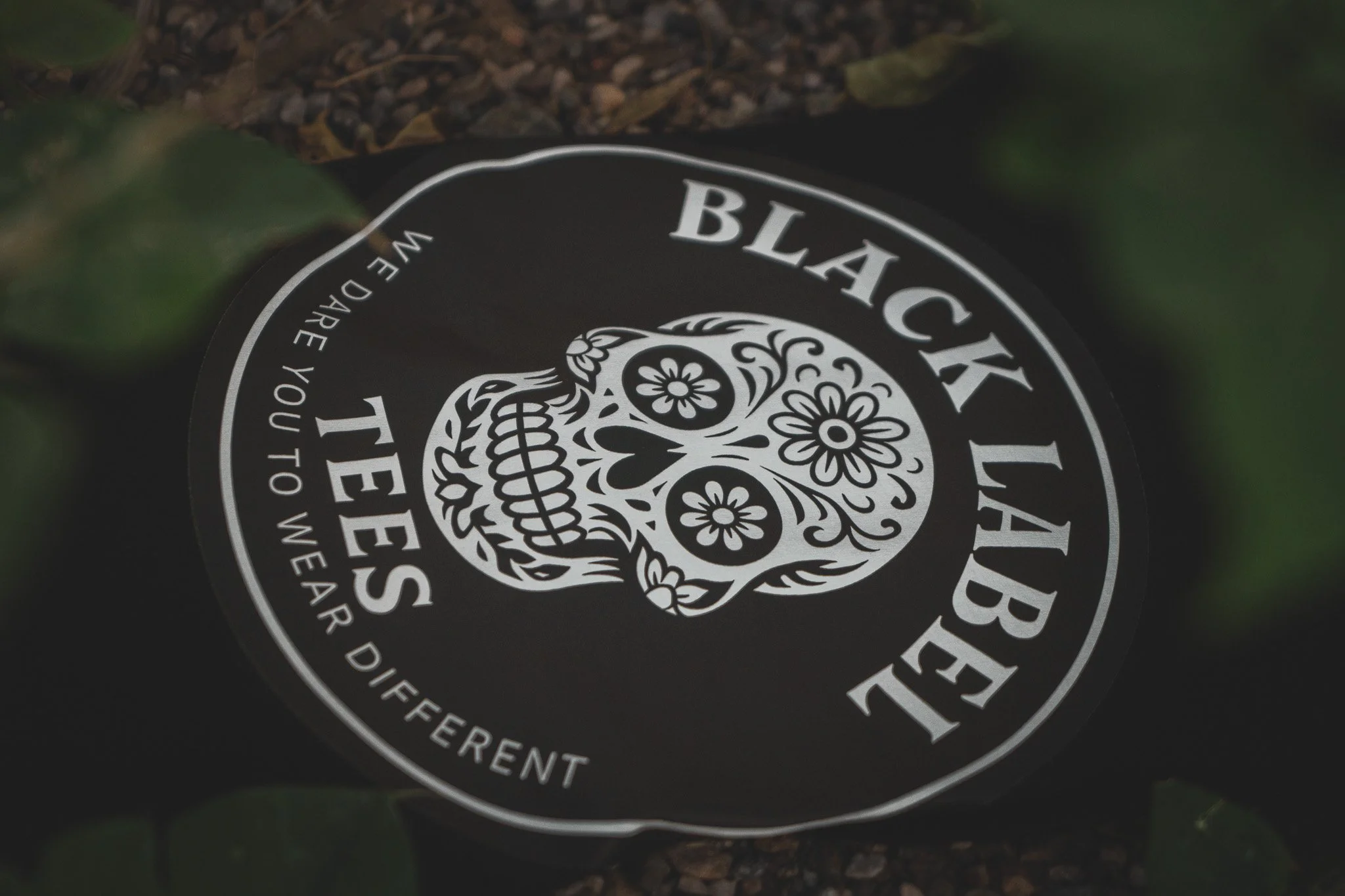 Close-up of a black circular sticker featuring a white skull decorated with floral patterns, with the text "Black Label Tees" and the phrase "We dare you to wear different."