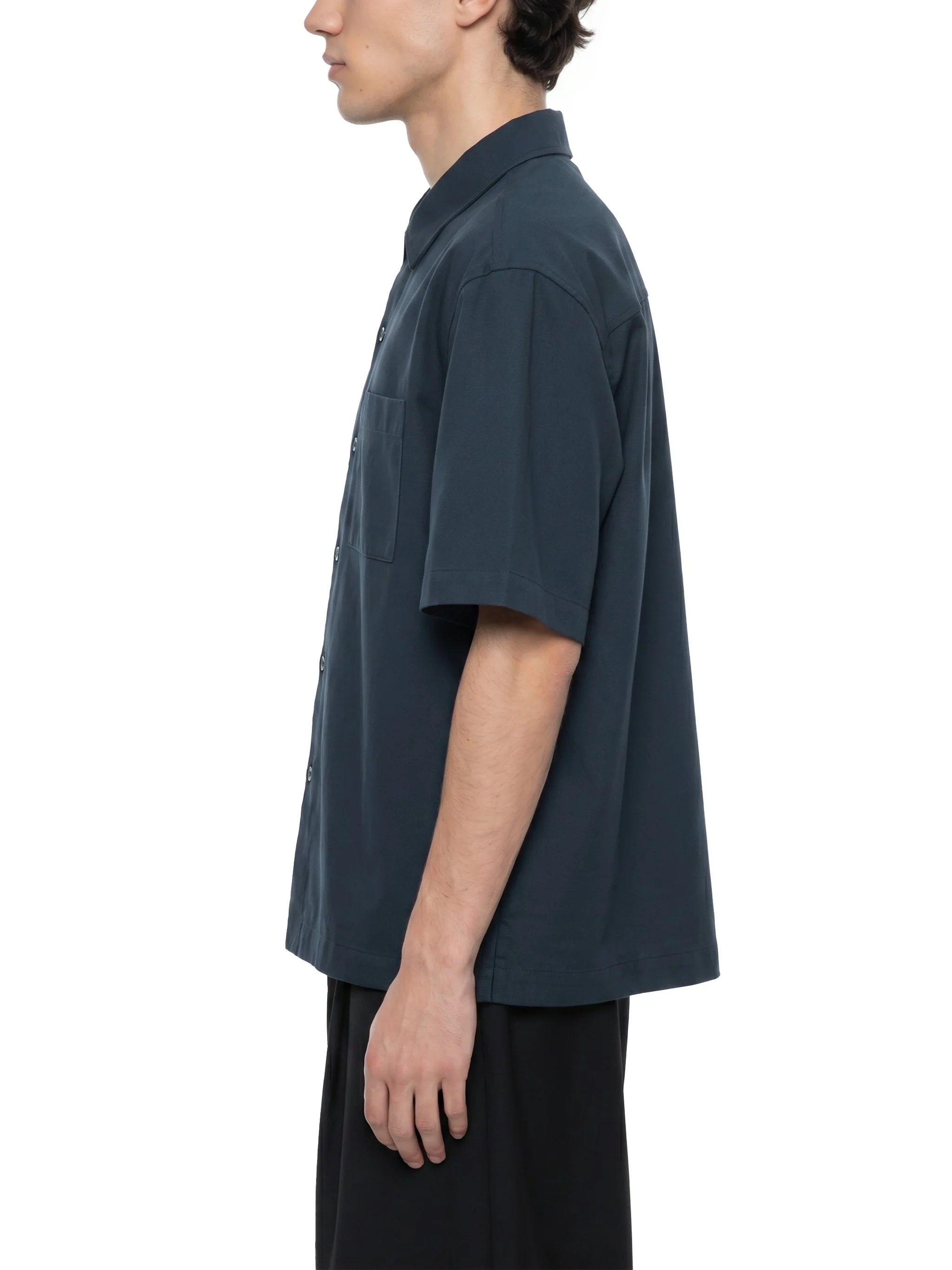 Side view of a young man wearing a dark blue short-sleeved button-up shirt, standing against a white background.