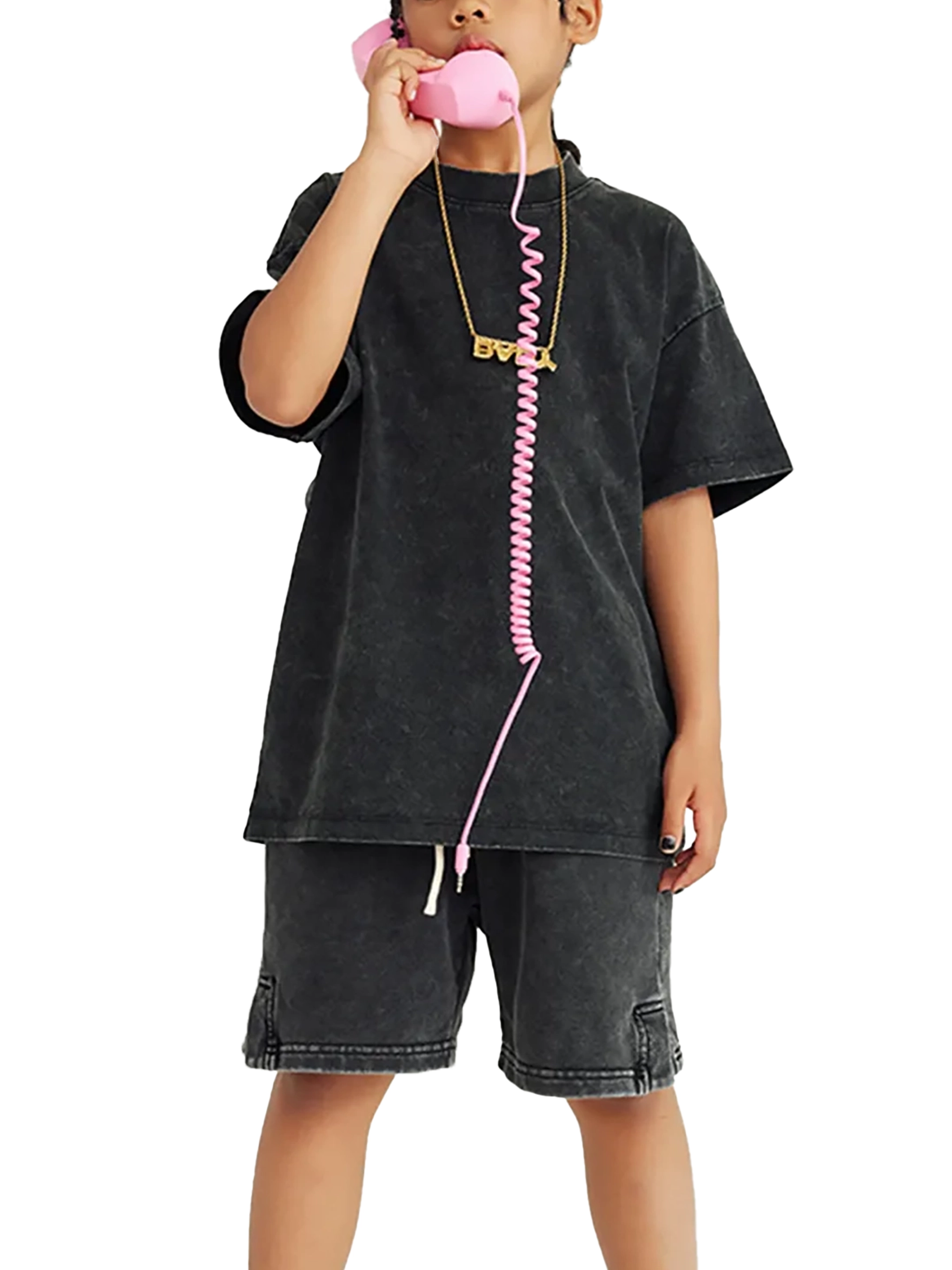 A child in gray shorts and a black t-shirt holding a pink retro-style telephone with a curly cord to their ear.