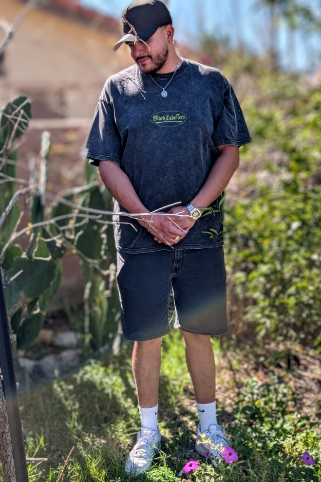A man standing outdoors among plants, wearing a black cap, dark graphic T-shirt, black shorts, white socks, and sneakers, with sunlight and greenery in the background.
