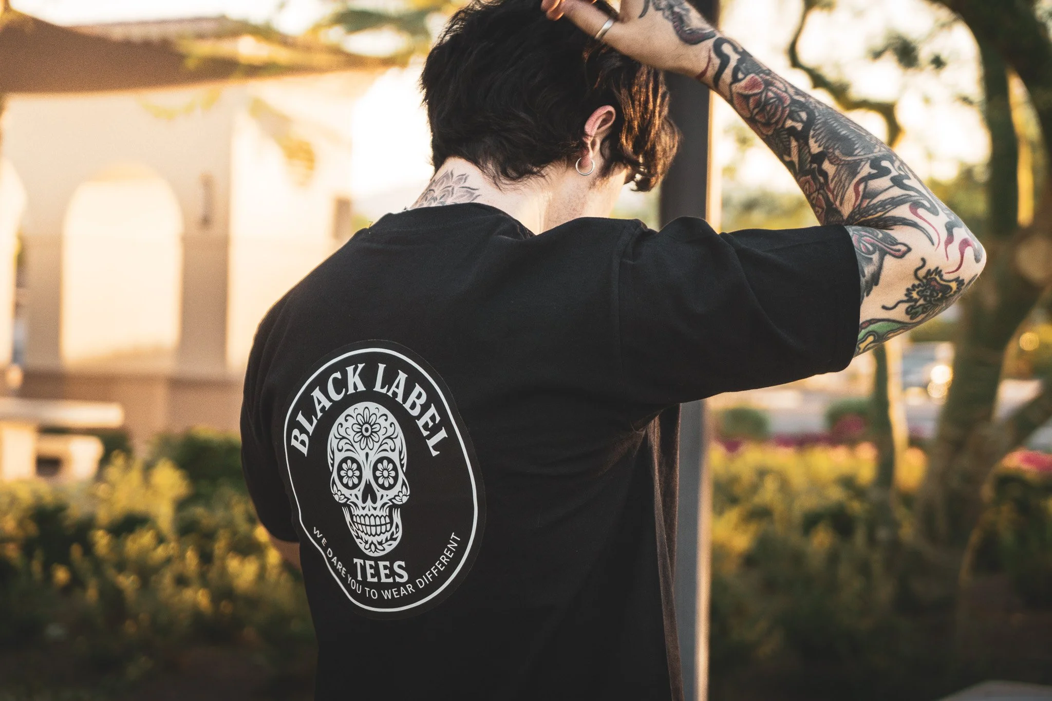 Back of a person wearing a black t-shirt with a white skull design and the words "Black Label Tees" on it, standing outside during sunset with tattoos visible on their arm.