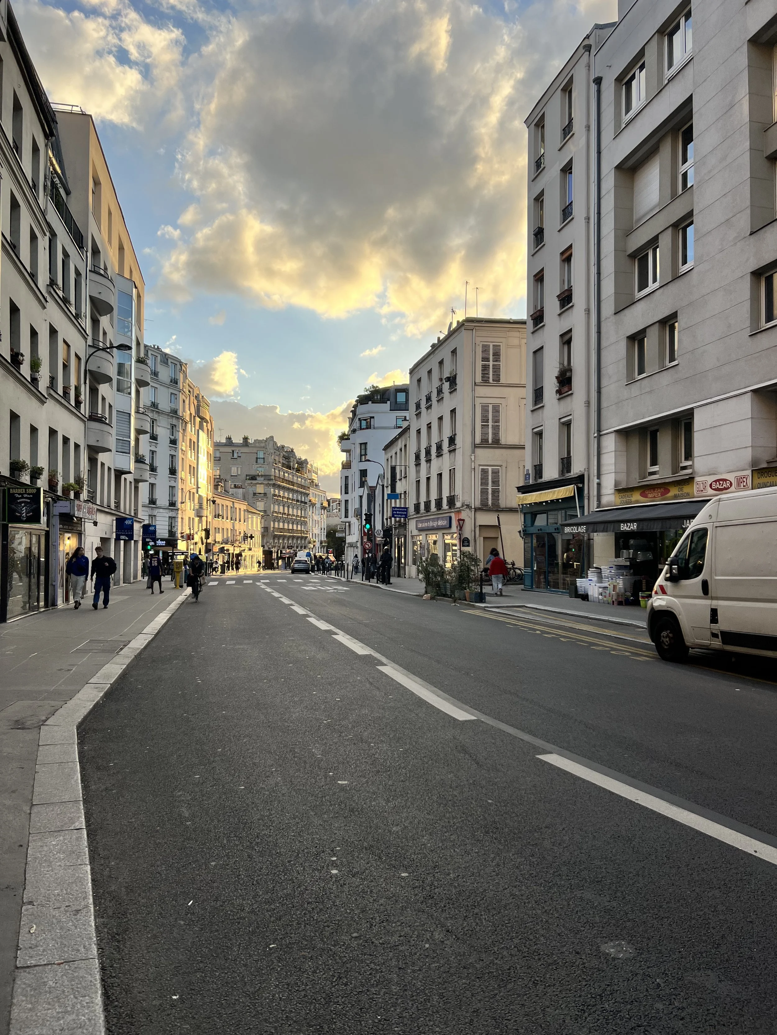 Rue de Bagnolet during sunset with pedestrians walking on the sidewalks in front of Quartier Rouge on a Sunday. Multi-story buildings with shops and apartments line the street. The sky has a mixture of clouds and sunlight.