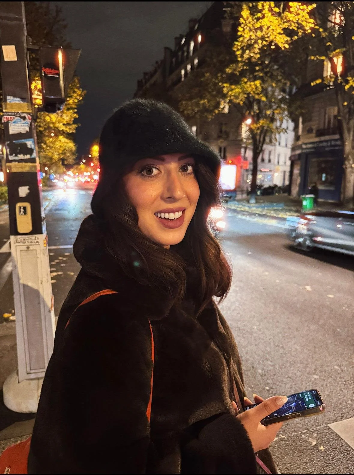 A young woman with a black hat and coat stands on a Paris street at night, smiling at the camera while holding a smartphone. The street behind her is illuminated with streetlights, and trees with yellow leaves line the sidewalk.