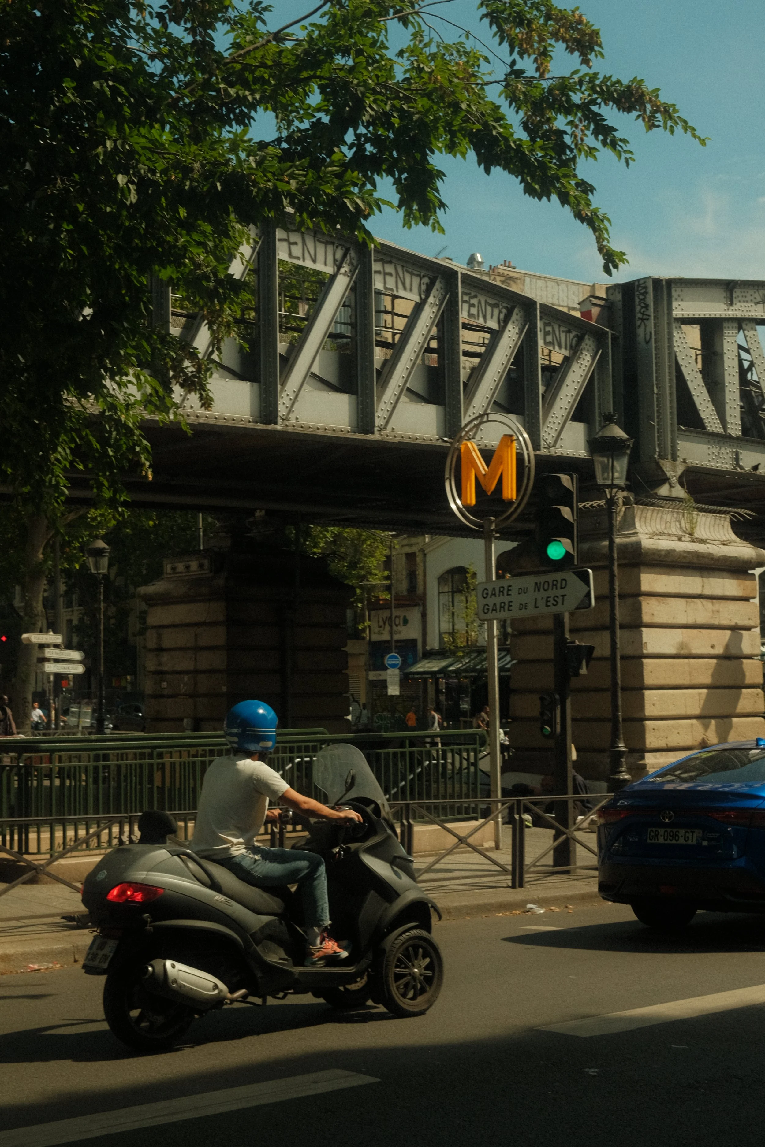 A Parisian street scene at showing a scooter rider at Barbès Rochechouart, with an orange 'M' metro sign, green traffic light, and signs indicating Gare du Nord and Gare de l'Est stations. The background includes shopfronts and street lamps.