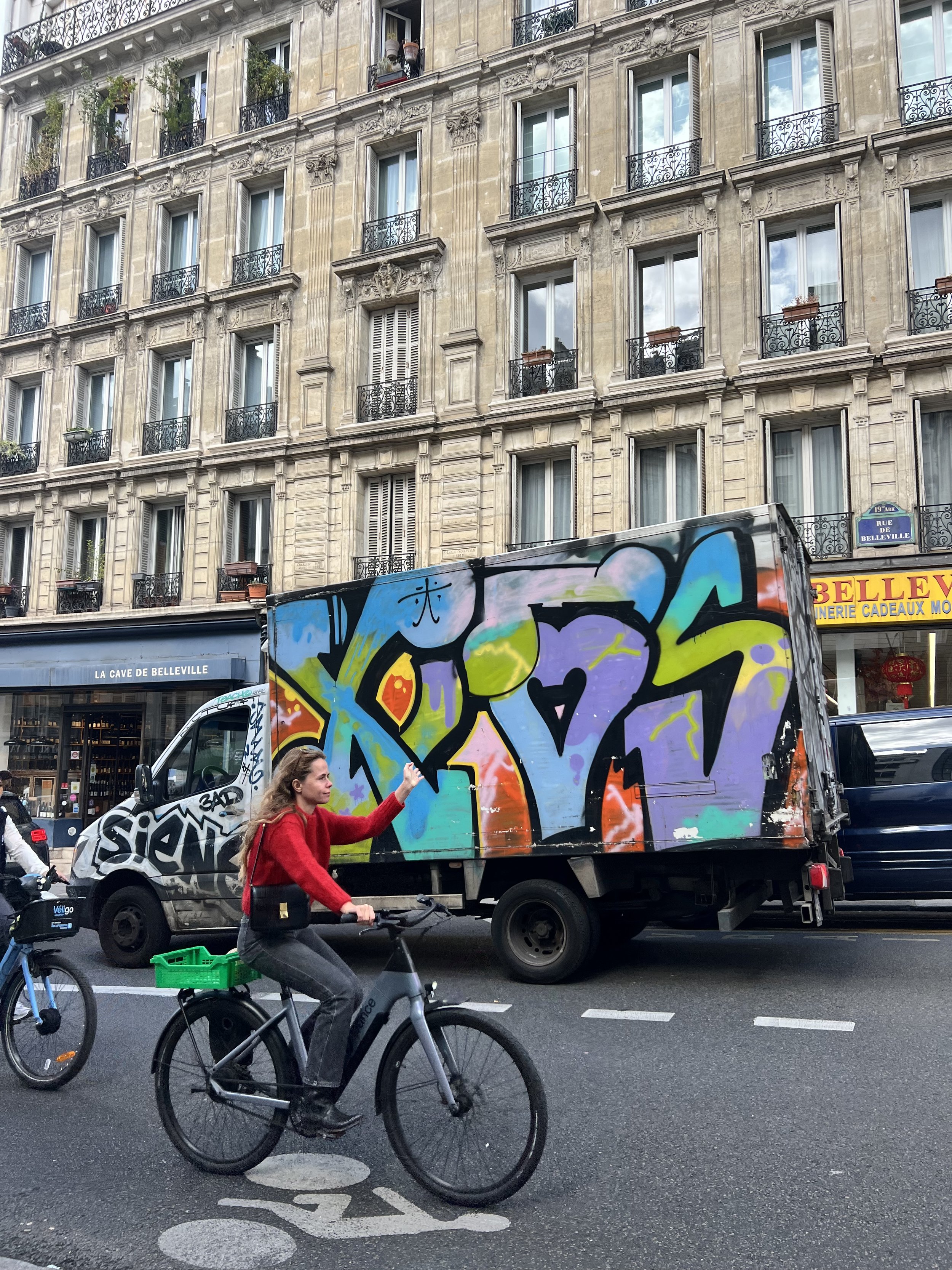 A woman in a red sweater riding a bicycle on a city street with a colorful graffiti-covered truck and Parisian apartment buildings in the background.