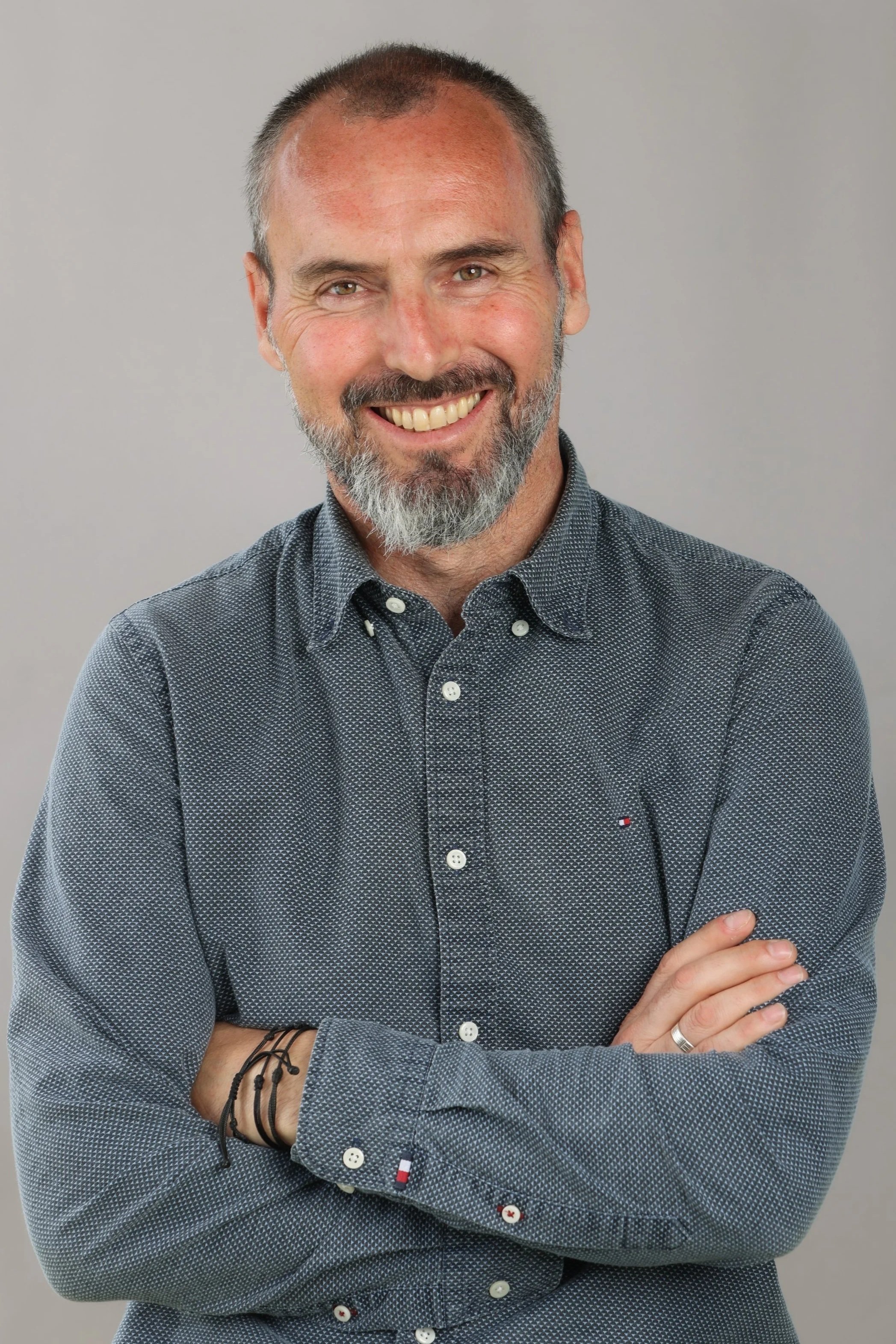 Homme souriant avec une barbe et des cheveux courts, portant une chemise à manches longues à petits motifs, le bras croisé. Fond gris clair.