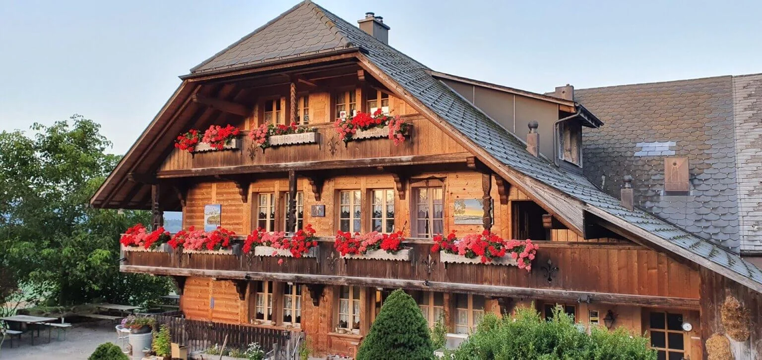 Unser traditionelles Berner Bauernhaus mit Balkon, Blumen in Blumenkästen und einem schrägen Dach, umgeben von Bäumen und Gärten. Ein Ort zum Wohlfühlen.