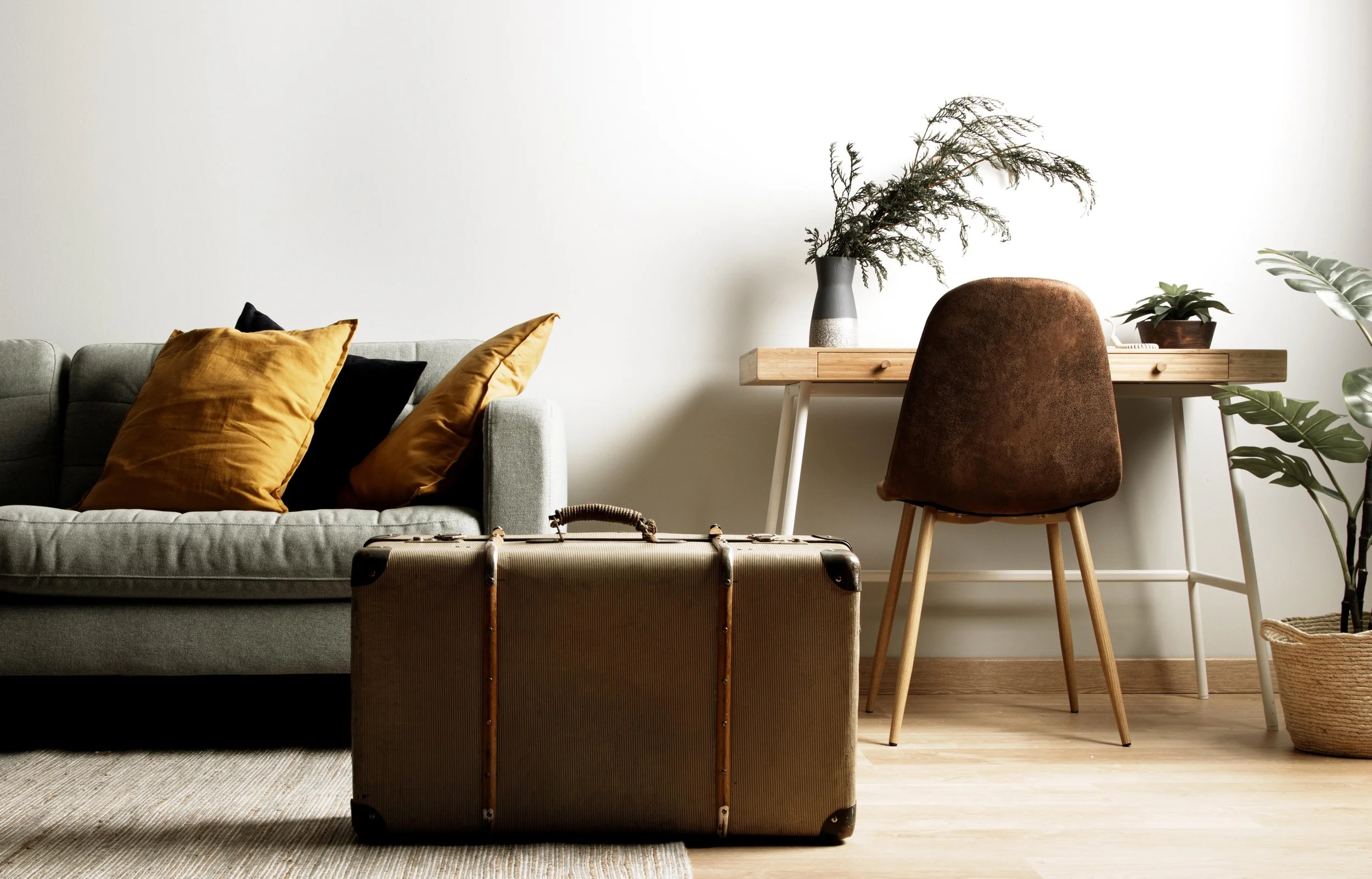 Living room with a light gray sofa with mustard and black pillows, a vintage suitcase in front, a wooden desk with plants, and a brown chair.